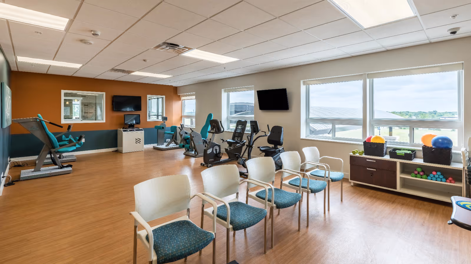 Bright fitness room with exercise bikes, resistance machines, a row of chairs, and shelves holding exercise balls and weights.