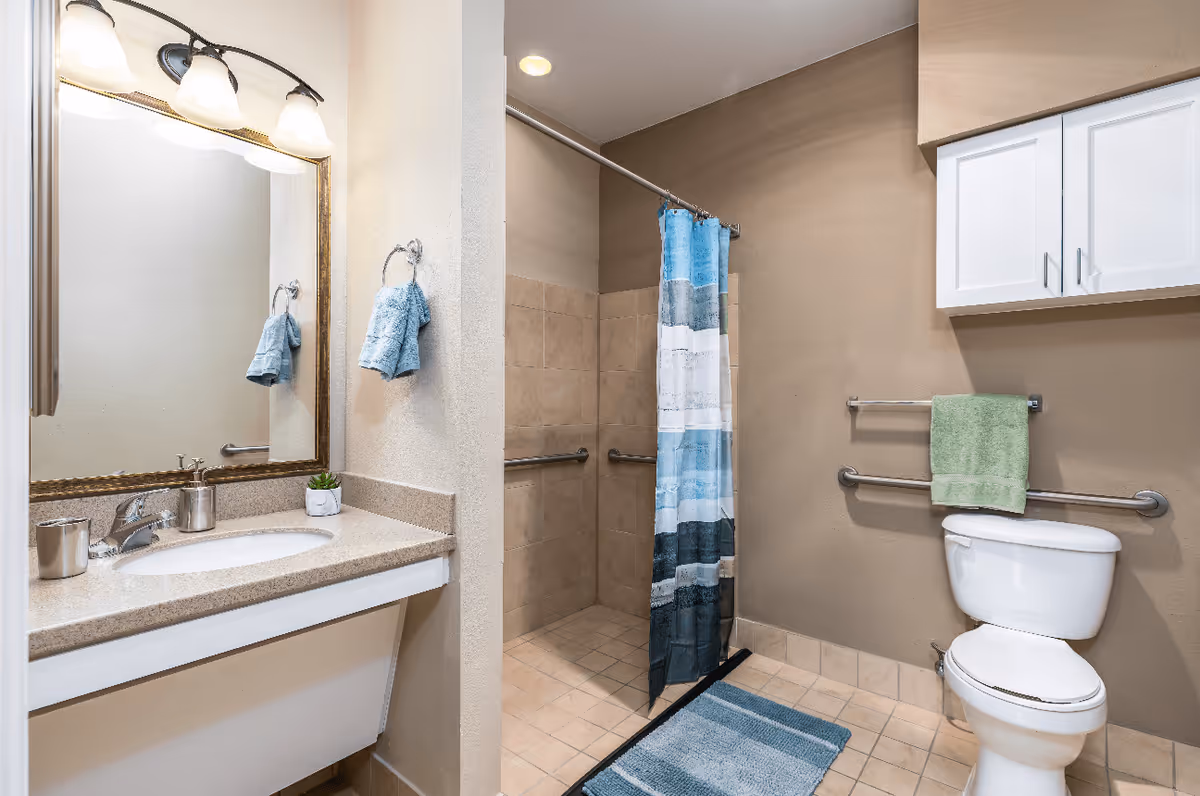 A clean and accessible bathroom featuring a sink with a countertop, a large mirror with three light fixtures above it, a towel ring with a blue towel, a walk-in shower with a blue and white striped shower curtain, grab bars on the shower walls, a toilet with a green towel hanging on a grab bar above it, and white cabinets mounted on the wall.