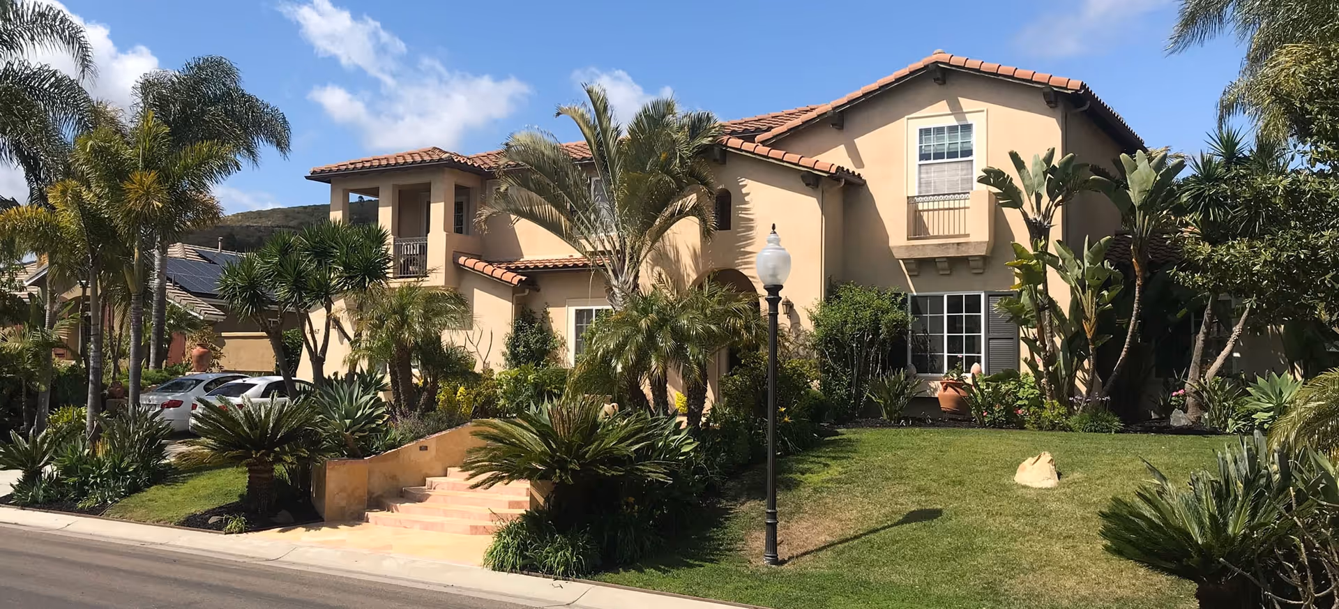 Two-story Mediterranean-style house with a red-tiled roof, palm trees, and a landscaped front lawn.