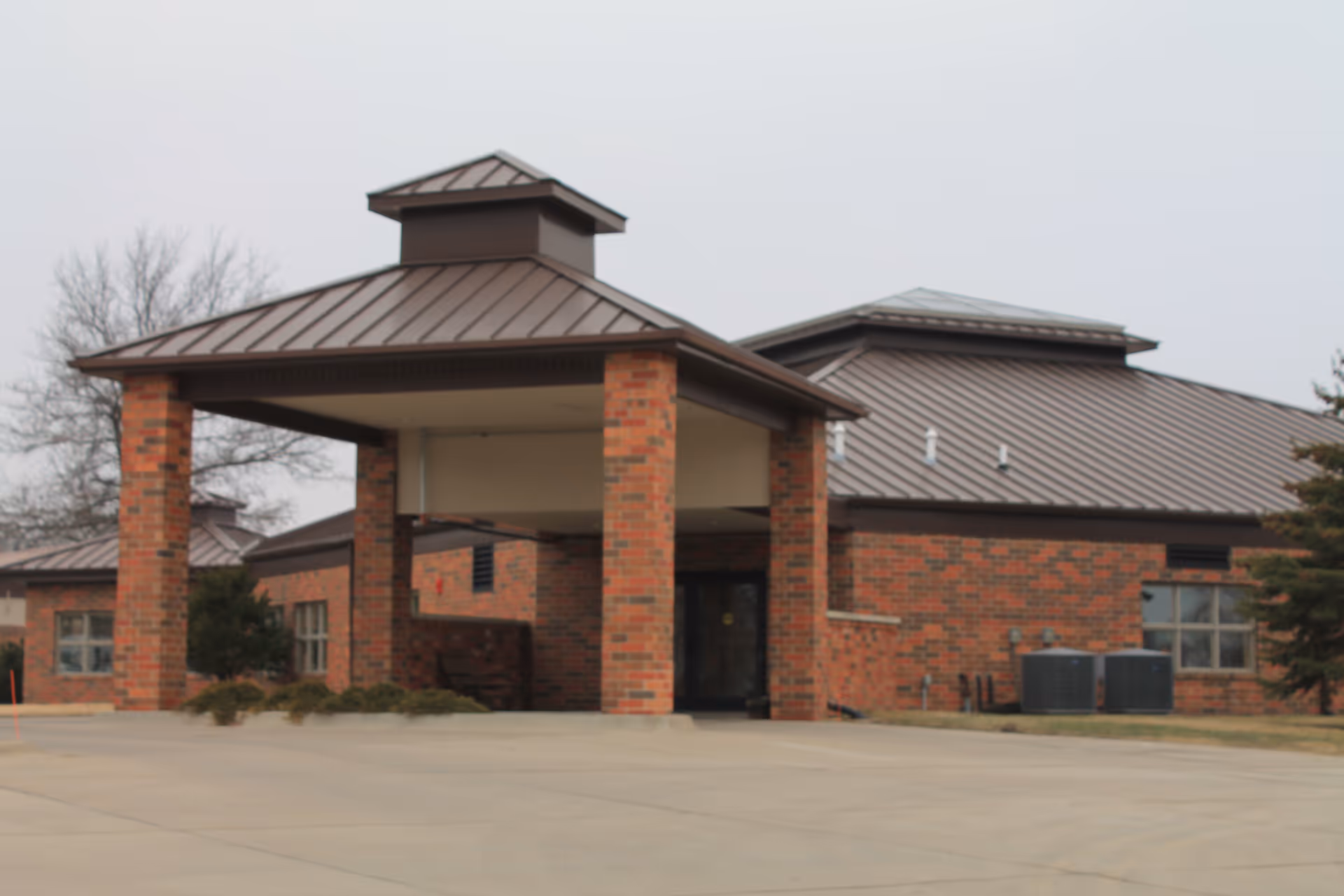 Exterior view of a brick building with a covered entrance supported by four brick columns and a metal roof, set against a cloudy sky with some trees visible in the background.