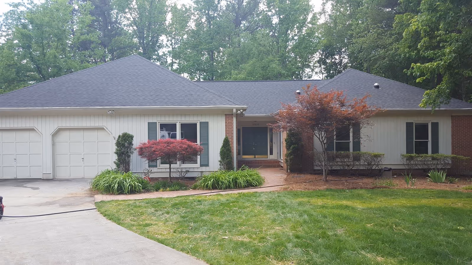 Front exterior view of a single-story residential building with a dark gray roof, white siding, and brick accents. The house has two garage doors on the left, a central entrance with a covered porch, and several windows with green shutters. The front yard features a well-maintained lawn, a curved concrete driveway, and landscaped areas with small trees and shrubs.