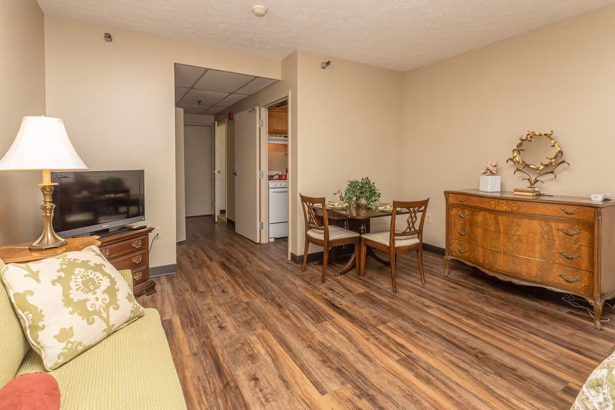 Interior view of a senior living facility room with wooden flooring, a green sofa with patterned cushions, a wooden TV stand with a flat-screen TV and a lamp, a small round dining table with two chairs and a plant centerpiece, and a wooden dresser with decorative items on top. A hallway and a small kitchen area with a stove are visible in the background.