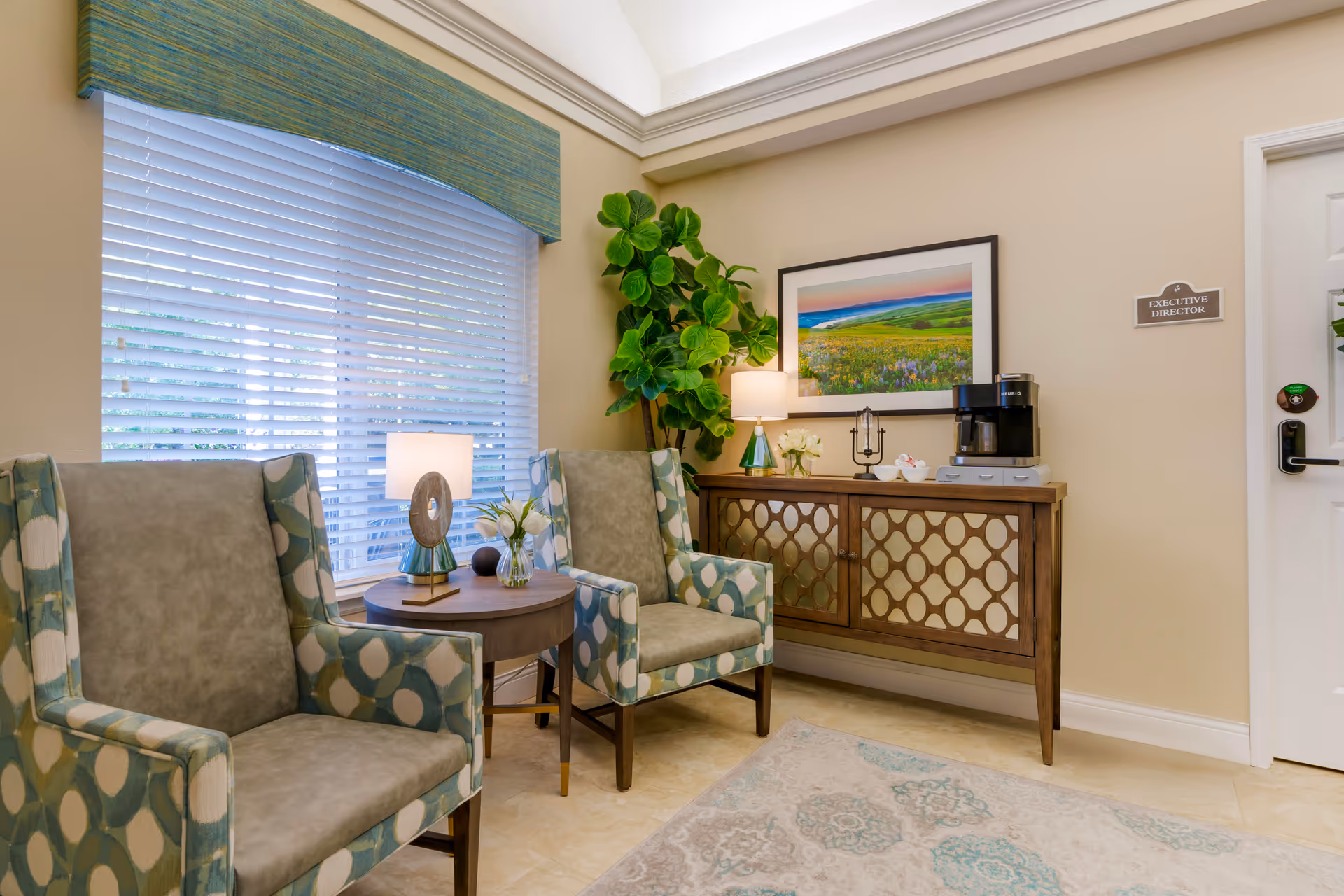 A cozy sitting area with two patterned armchairs and a small round wooden table between them, holding a decorative lamp and a small vase with flowers. Behind the chairs is a large window with white blinds and a green valance. To the right, there is a wooden sideboard with a geometric pattern on the doors, topped with a lamp, a coffee maker, and decorative items. A framed landscape painting hangs above the sideboard, and a door labeled 'Executive Director' is visible on the right side of the image. A large green potted plant is placed in the corner between the window and the sideboard.