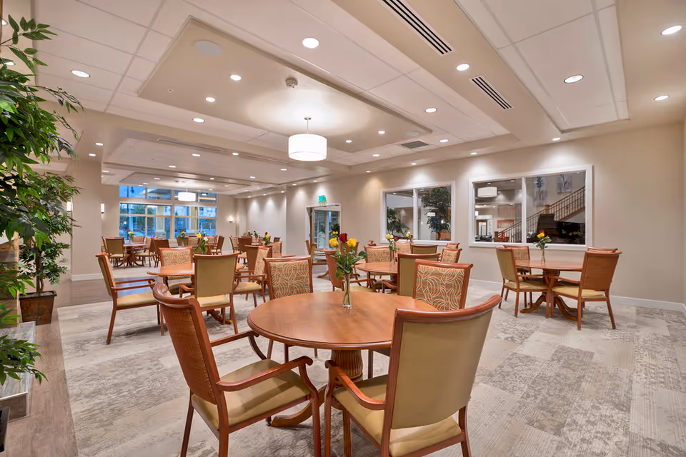 A spacious and well-lit dining room in a senior living facility with multiple round wooden tables, each surrounded by cushioned chairs. Small vases with flowers are placed on each table. The room features large windows, recessed lighting, and neutral-colored walls and carpet.