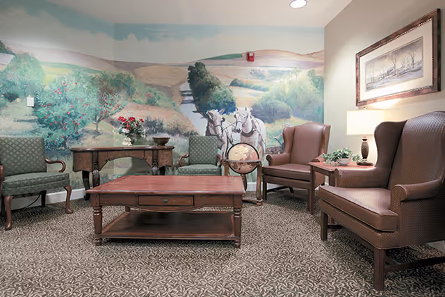 A cozy sitting area with two brown leather armchairs, two green patterned chairs, a wooden coffee table, and a side table with a lamp and plant. The back wall features a large mural depicting a rural landscape with trees, hills, and two white horses pulling a wagon. There is also a framed picture on the right wall.