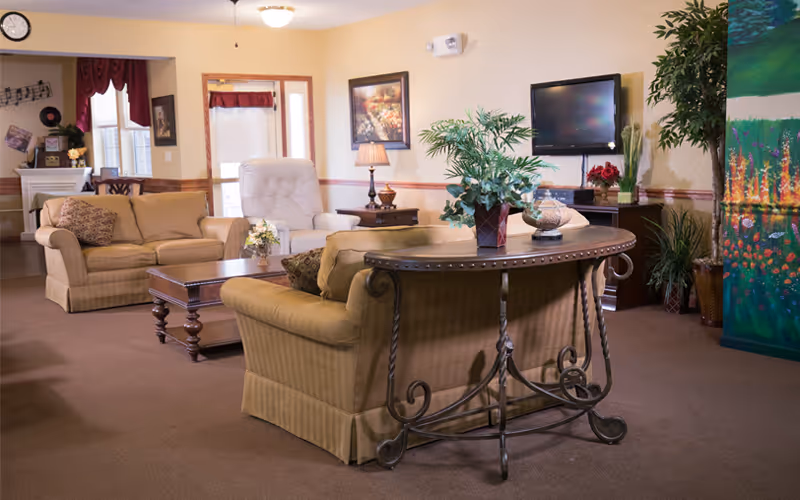 Comfortable communal living room with upholstered sofas, coffee table, wall-mounted TV, plants, and decorative furnishings.