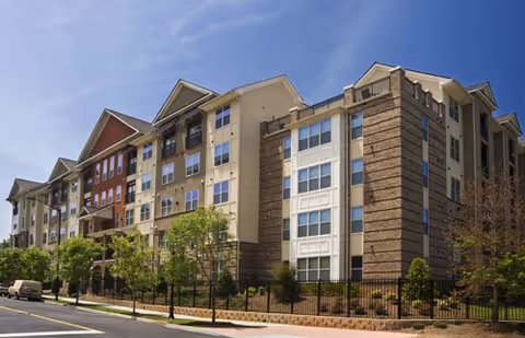 Exterior view of a multi-story senior living facility building with a combination of brick and beige siding, multiple windows, and a fenced landscaped area with trees and shrubs along the sidewalk under a clear blue sky.