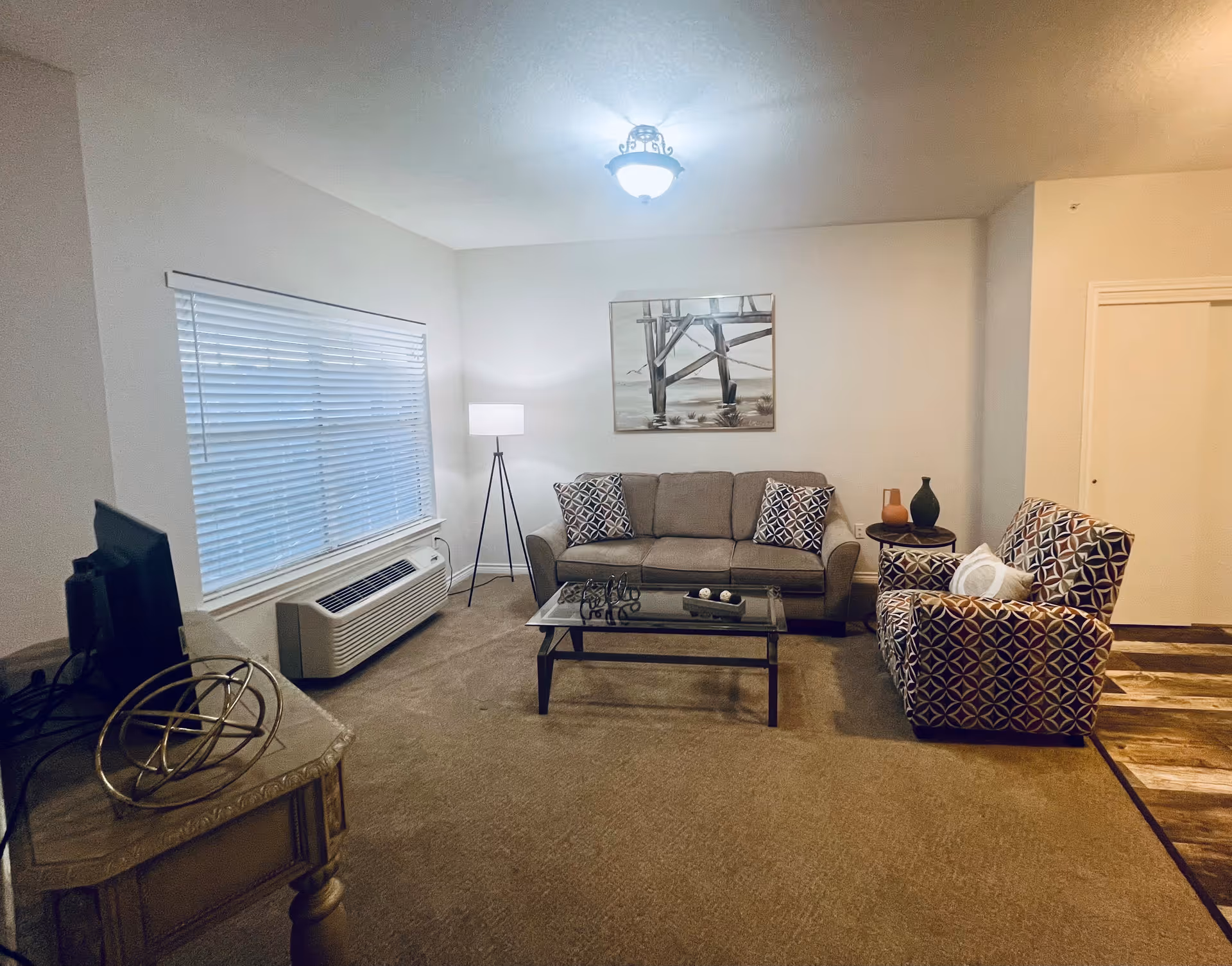 A cozy living room with a gray sofa adorned with patterned pillows, a matching patterned armchair with a white pillow, a glass coffee table, and a side table with decorative vases. A floor lamp stands near a window with closed blinds, and a wall painting depicting a pier over water hangs above the sofa. The room has beige carpet and a ceiling light fixture.