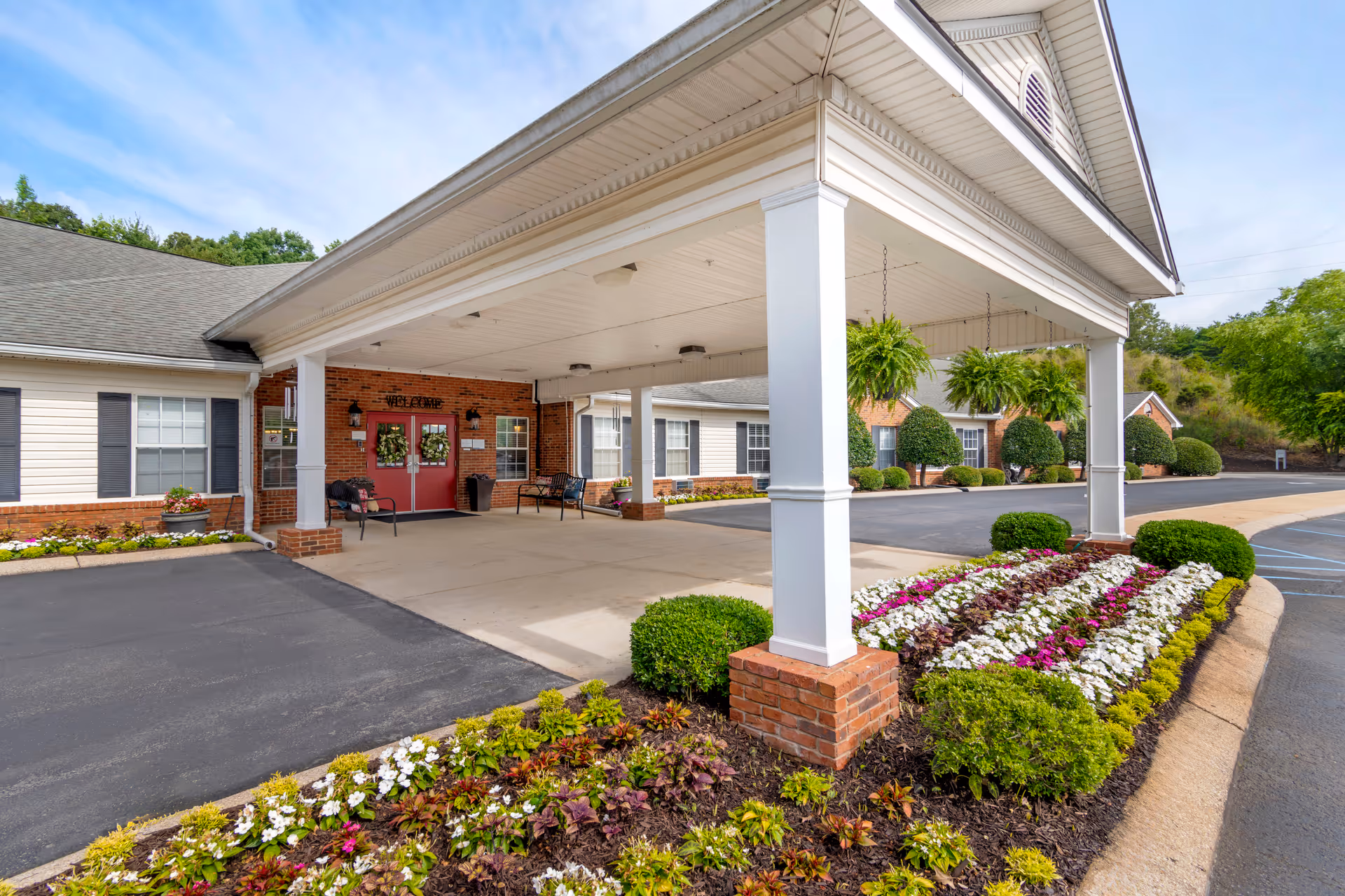 Entrance of Brookdale Hixson facility featuring a covered drop-off area with white pillars and hanging green plants. The building has red brick and white siding with black shutters. There are benches near the red double doors with wreaths and a 'WELCOME' sign above them. The surrounding area includes neatly landscaped flower beds and bushes.