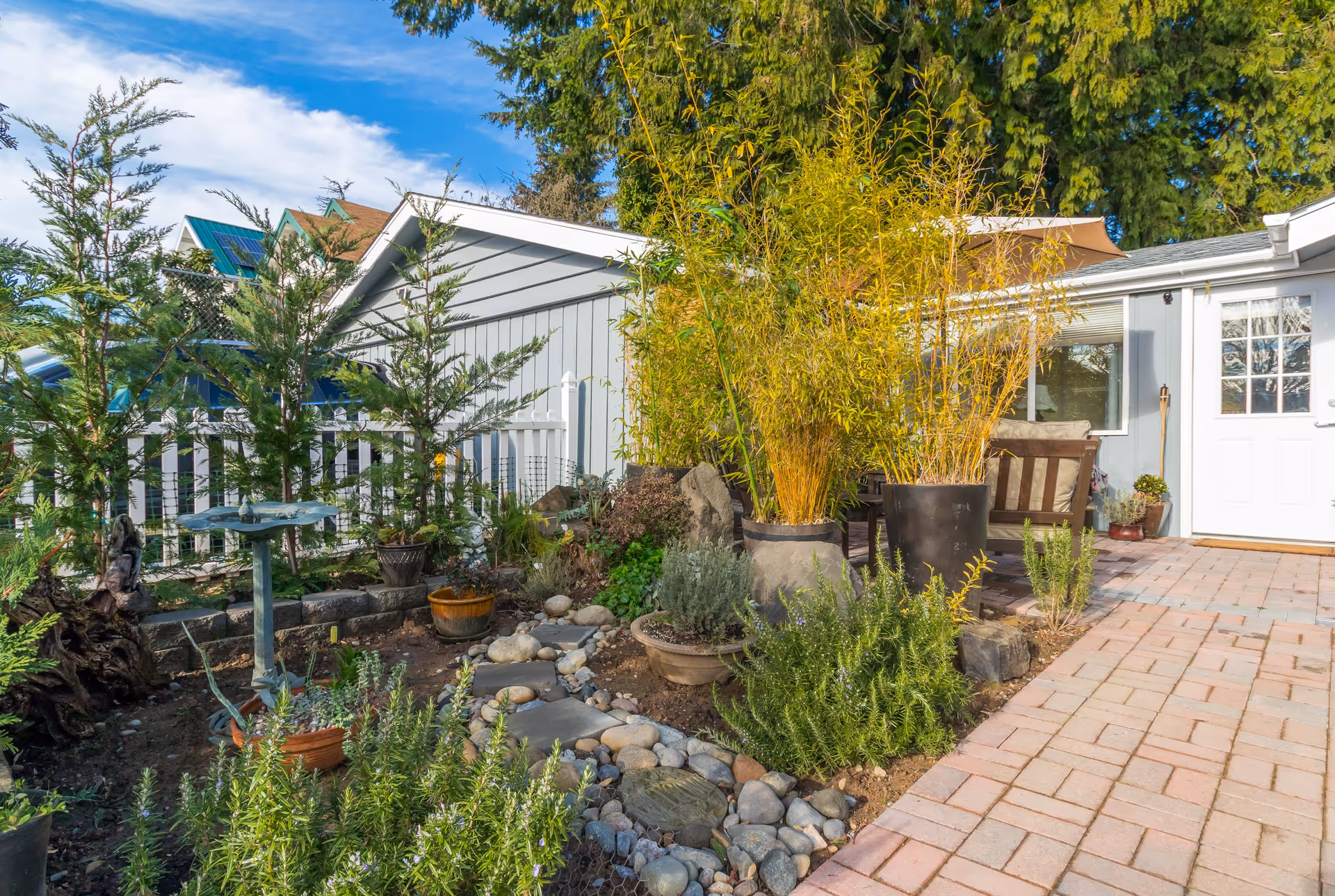 Small garden patio beside a light-gray house with potted bamboo, shrubs, a birdbath and a brick walkway leading to a white door.