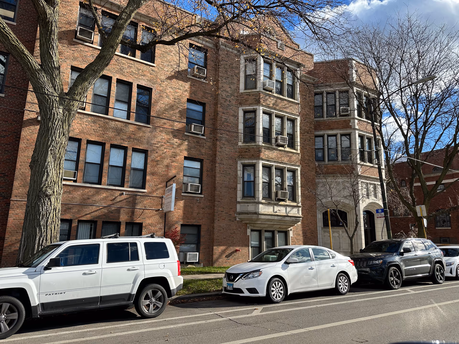 Exterior view of a multi-story brick building with several windows, some with air conditioning units. There are three parked cars along the street in front of the building, and leafless trees are visible. The sky is partly cloudy with blue patches.