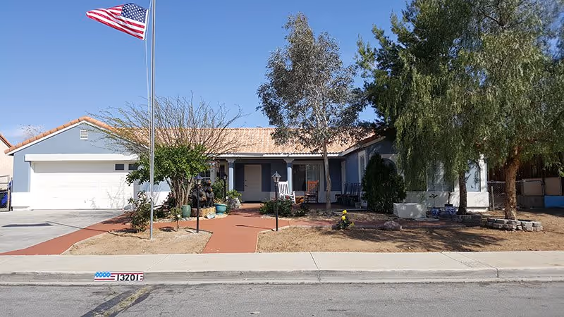 Single-story residential building with a beige tiled roof and blue-gray exterior walls. The front yard has a paved walkway, some trees, and an American flag on a tall flagpole. There is a driveway leading to a white garage door on the left side of the house.