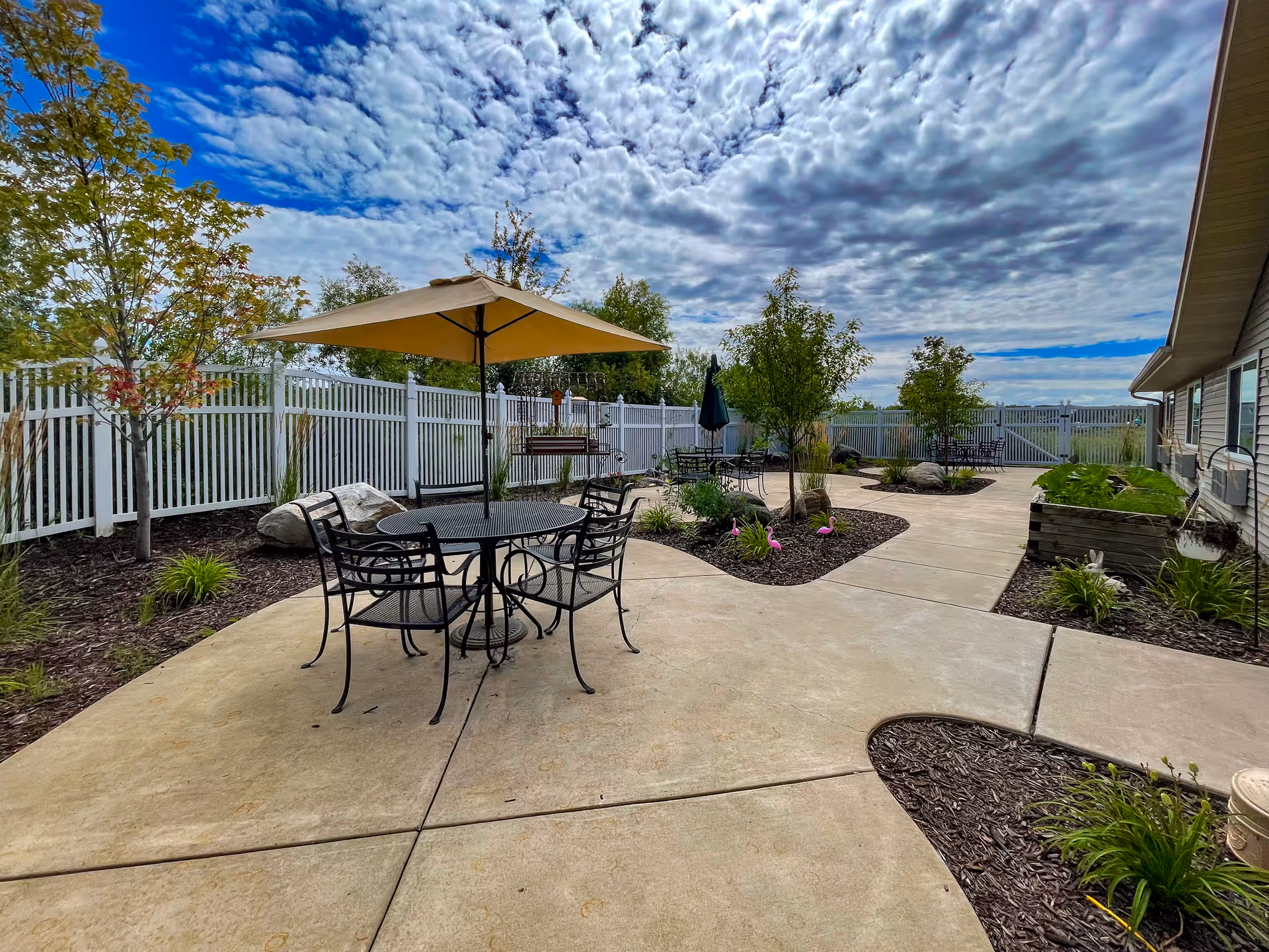 Outdoor patio area with metal tables and chairs, some shaded by large umbrellas, surrounded by landscaped garden beds with small trees and plants, enclosed by a white fence under a partly cloudy sky.