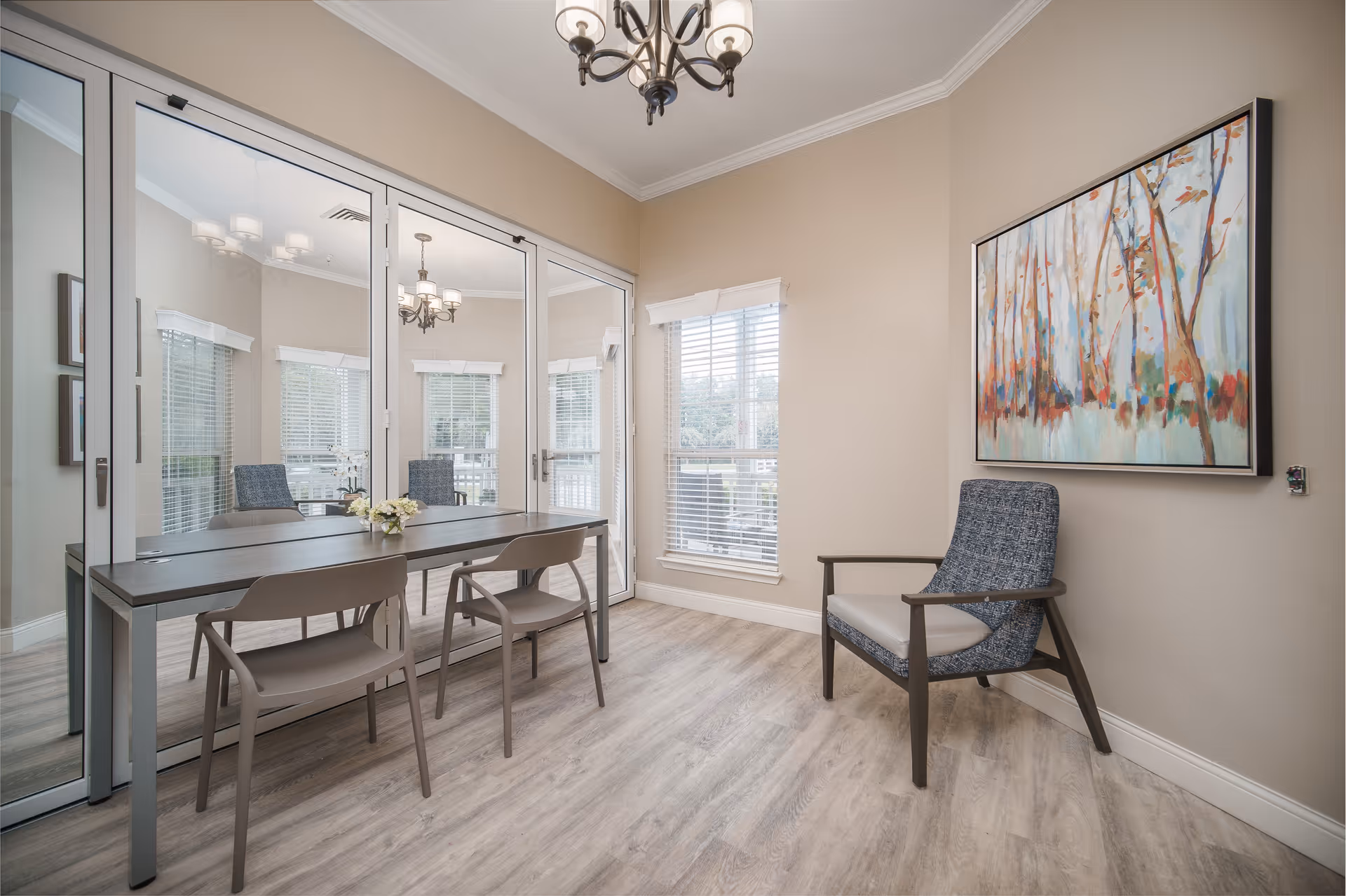 A bright and modern interior room with light wood flooring, beige walls, and a large window with white blinds. There is a rectangular table with two chairs on one side and a single upholstered armchair with a blue patterned cushion on the other side. A colorful abstract painting of trees hangs on the wall above the armchair. A chandelier with multiple lights hangs from the ceiling, and a large mirror covers one wall, reflecting the room.
