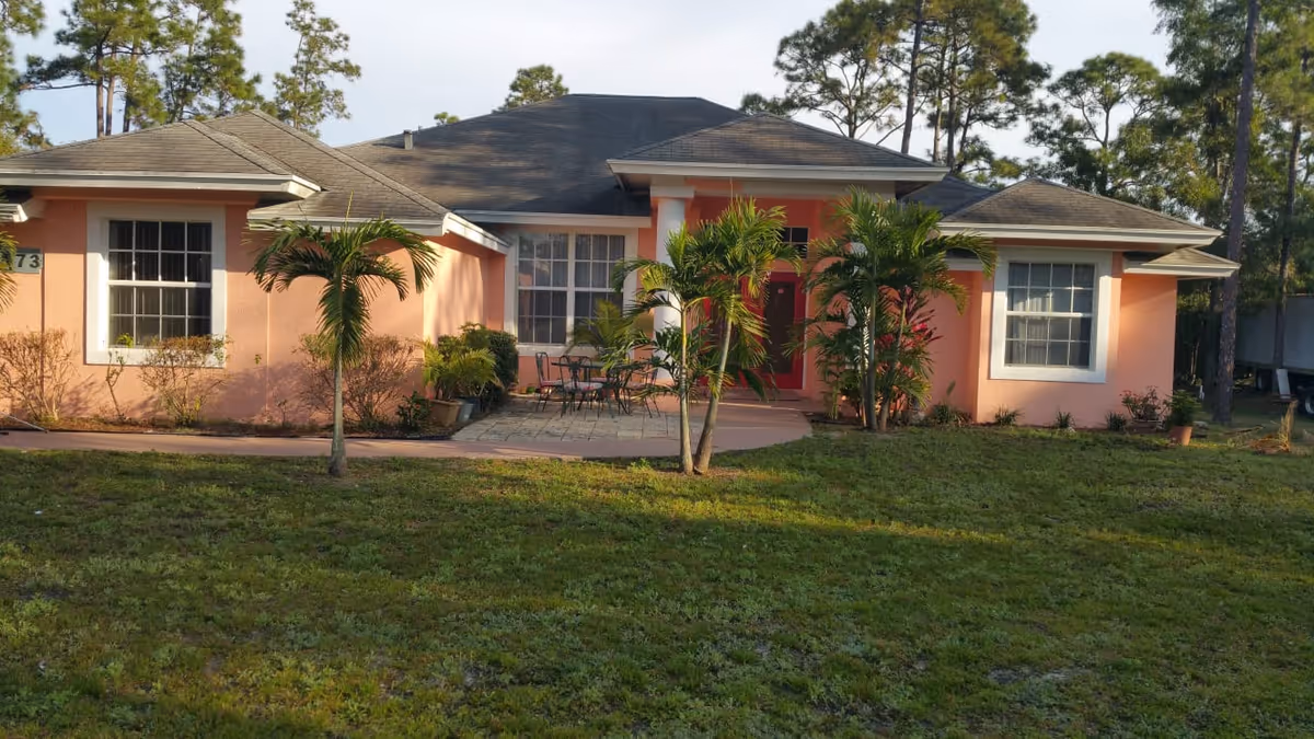 Front exterior view of a single-story pink house with a dark roof, two large windows, a red front door, and a small patio area with outdoor furniture. There are several palm trees and shrubs in the front yard, and a grassy lawn extends in front of the house.