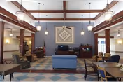 Spacious senior community common area with high ceilings and exposed wooden beams. The room features a blue couch in the center, several armchairs, tables with chairs, and a fireplace on the left side. Pendant lights hang from the ceiling, and a large framed artwork is displayed on the far wall above a cabinet with a television.