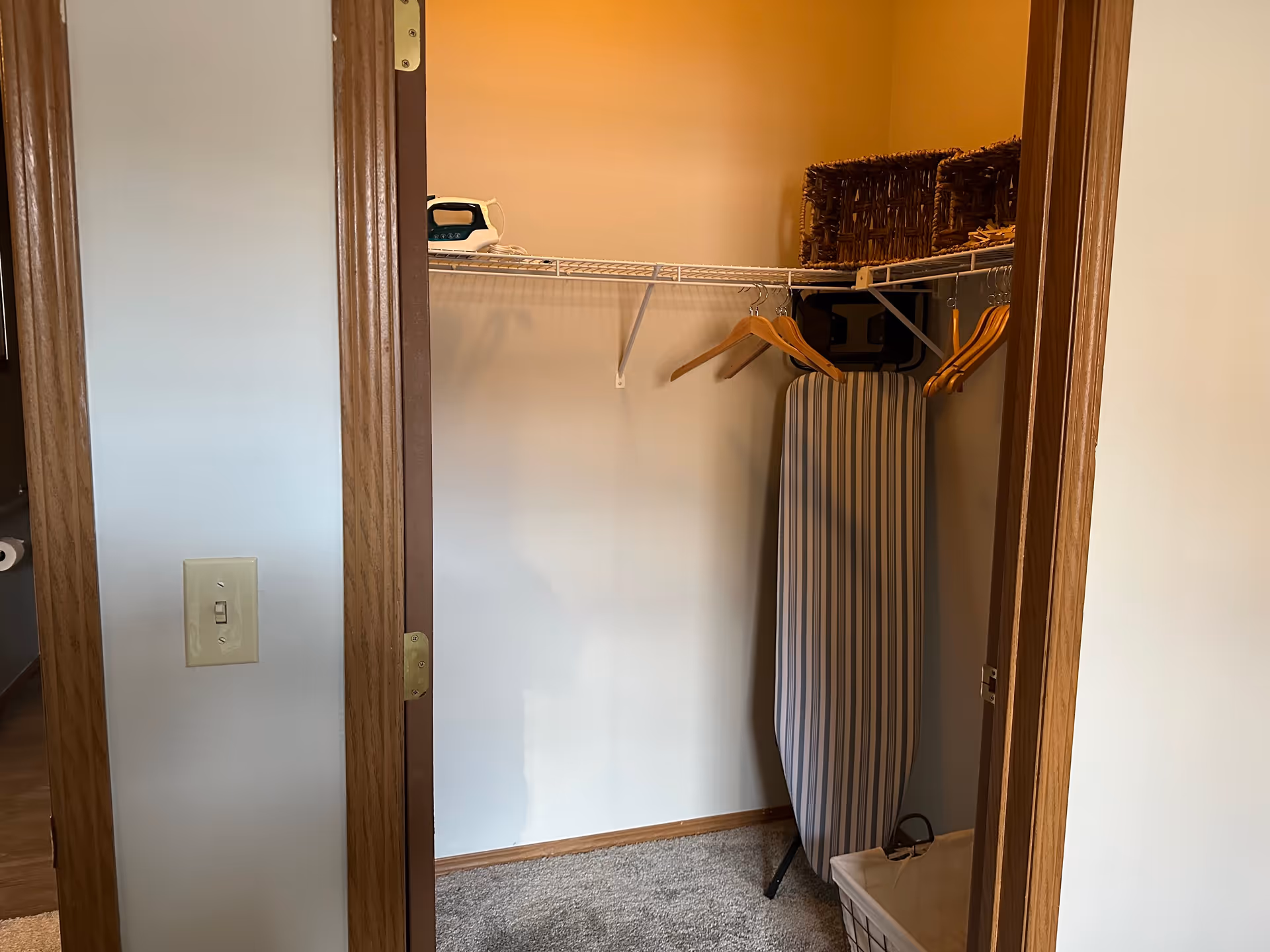 A small closet with a wire shelf holding an iron and woven baskets. Several wooden hangers hang from the shelf. An ironing board is standing upright against the wall, and a laundry basket is on the floor. The closet has beige walls and carpeted floor, with wooden door frames.