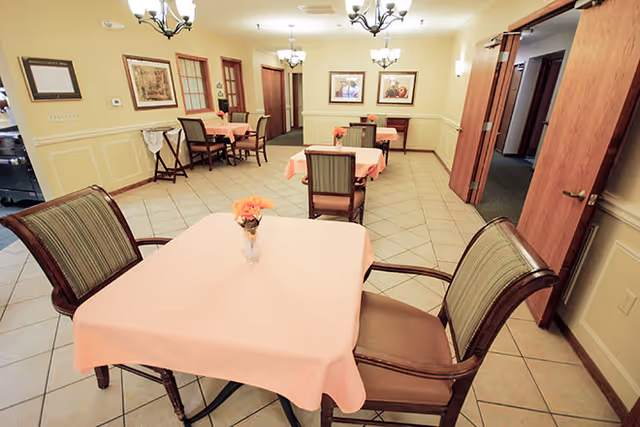 A dining room with several tables covered in pink tablecloths, each with a small flower arrangement. The room has tiled floors, beige walls with white wainscoting, framed artwork, and multiple chandeliers providing warm lighting. Wooden doors and windows are visible along the walls.