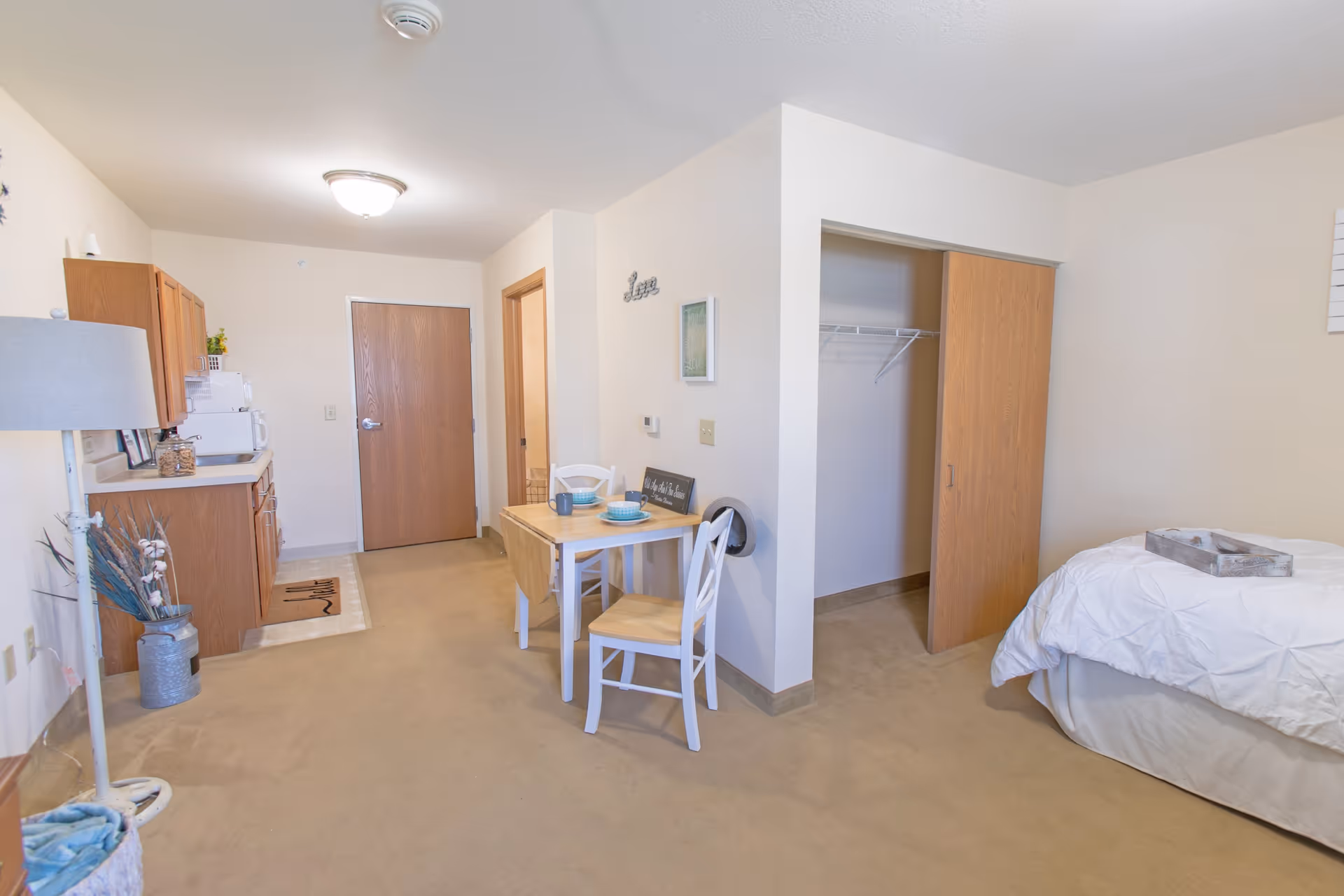Interior view of a senior living studio apartment featuring a small kitchenette with wooden cabinets and white appliances, a small dining table set with two chairs, an open closet with sliding wooden doors, and a bed with white bedding. The room has beige carpet and light-colored walls with minimal decor.