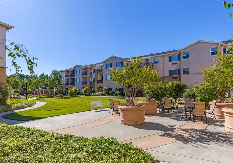Outdoor courtyard area of Heritage Estate Senior Apartments featuring a paved walkway, large potted plants, several patio tables with chairs, a white bench, green grass, and a three-story residential building in the background under a clear blue sky.