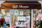 Interior gift shop with a brown awning reading 'Gift Shoppe' and shelves of flowers, cards, and gifts visible through the window.