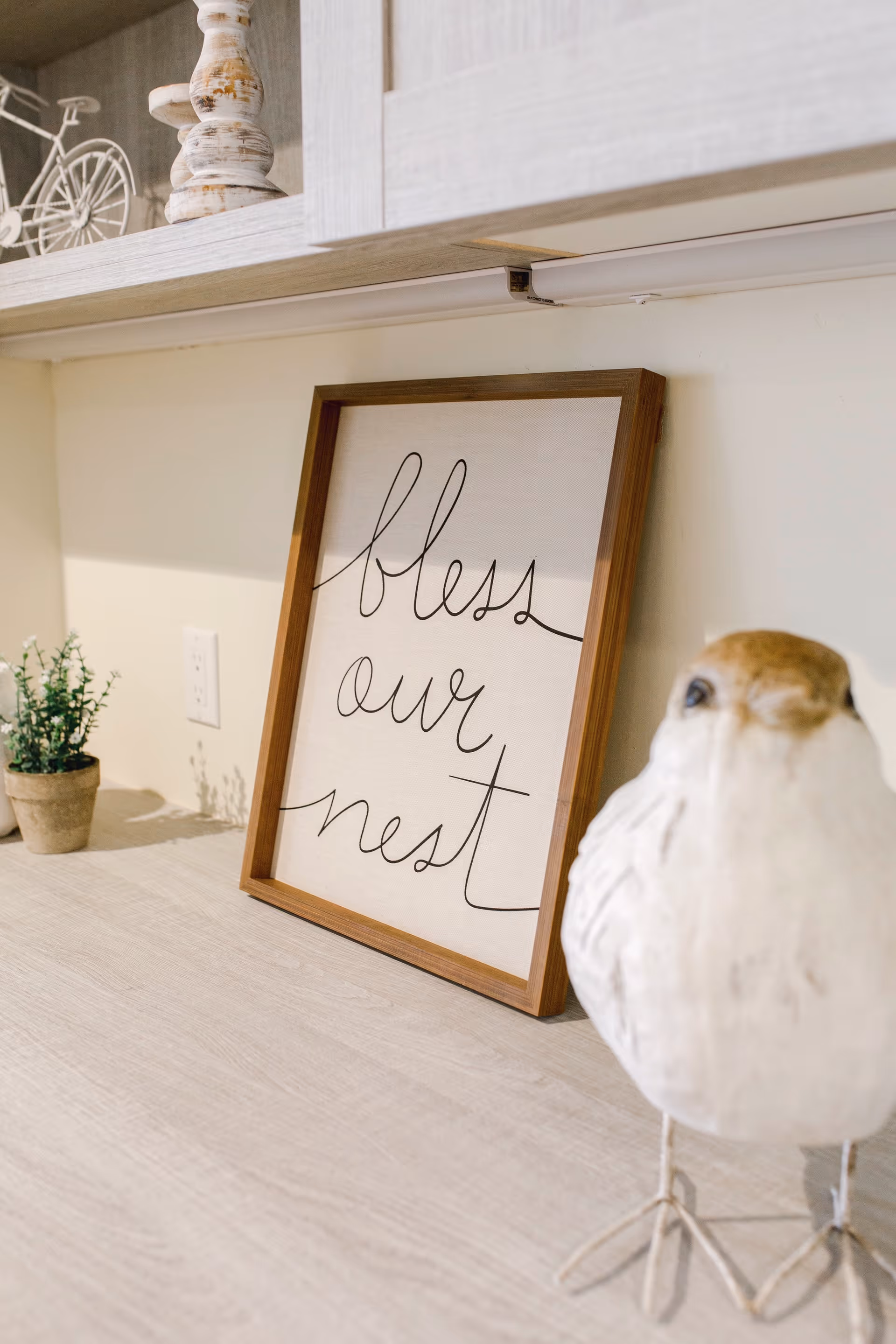 Framed sign reading "bless our nest" leaning on a light wood countertop with a decorative bird sculpture and small potted plant under cabinets.
