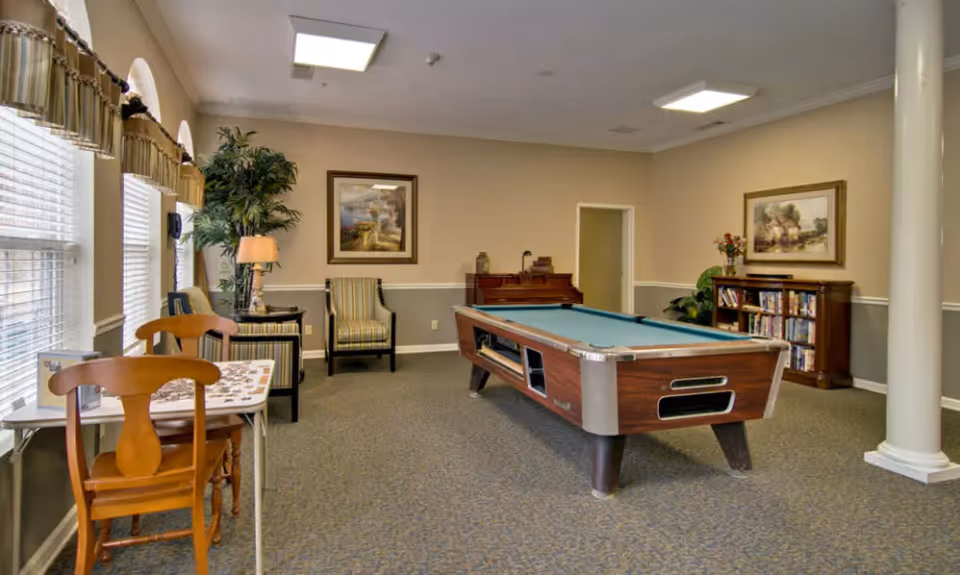 A recreational room with a pool table in the center, a small table with two wooden chairs on the left, two armchairs near the back wall, a piano, a bookshelf filled with books, framed paintings on the walls, a tall potted plant, and large windows with valances letting in natural light.