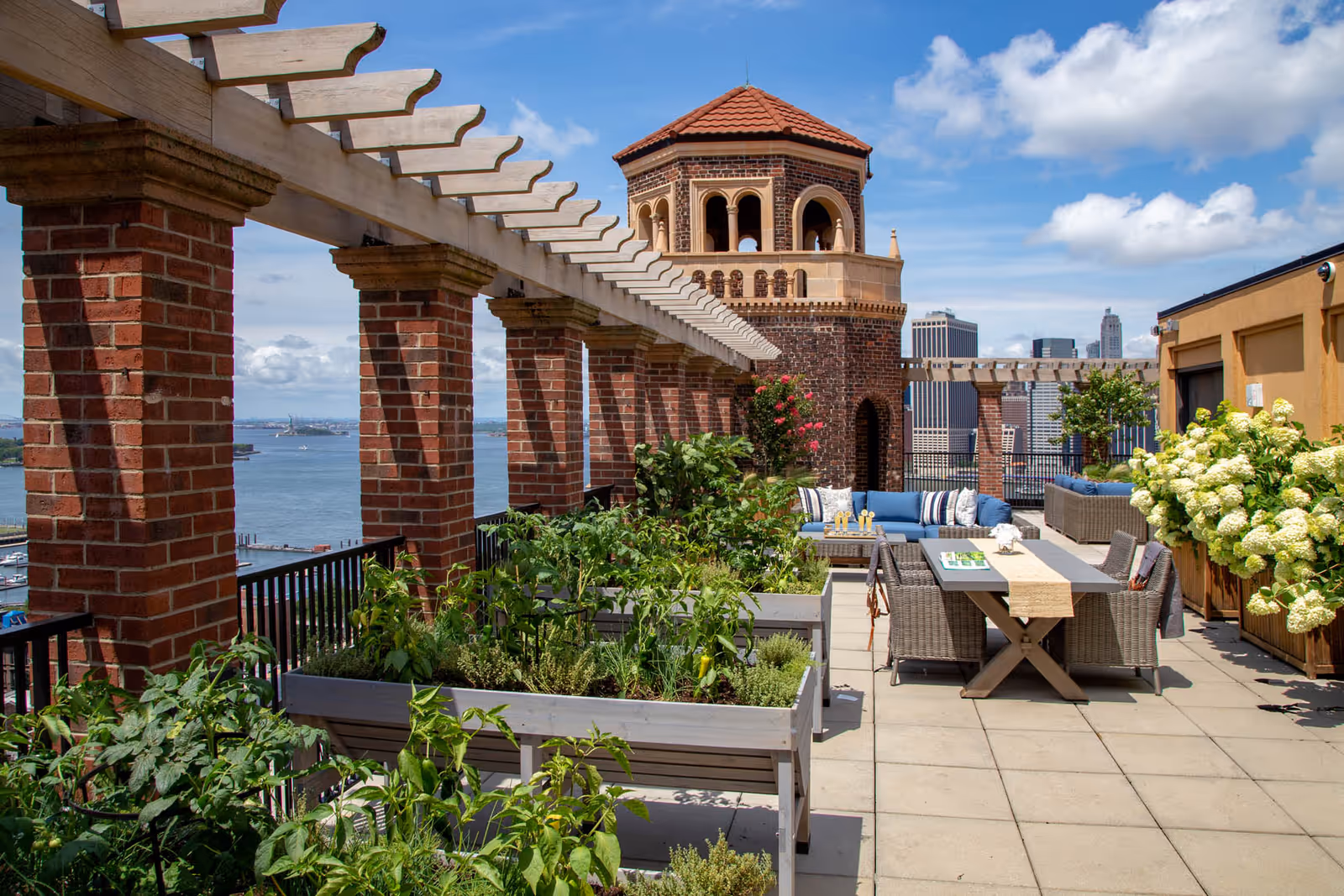 Outdoor rooftop garden area with raised planter boxes filled with green plants and herbs, a pergola with brick columns, a seating area with blue cushioned sofas, a dining table with wicker chairs, and a brick tower structure in the background under a partly cloudy sky with a city skyline and water view.
