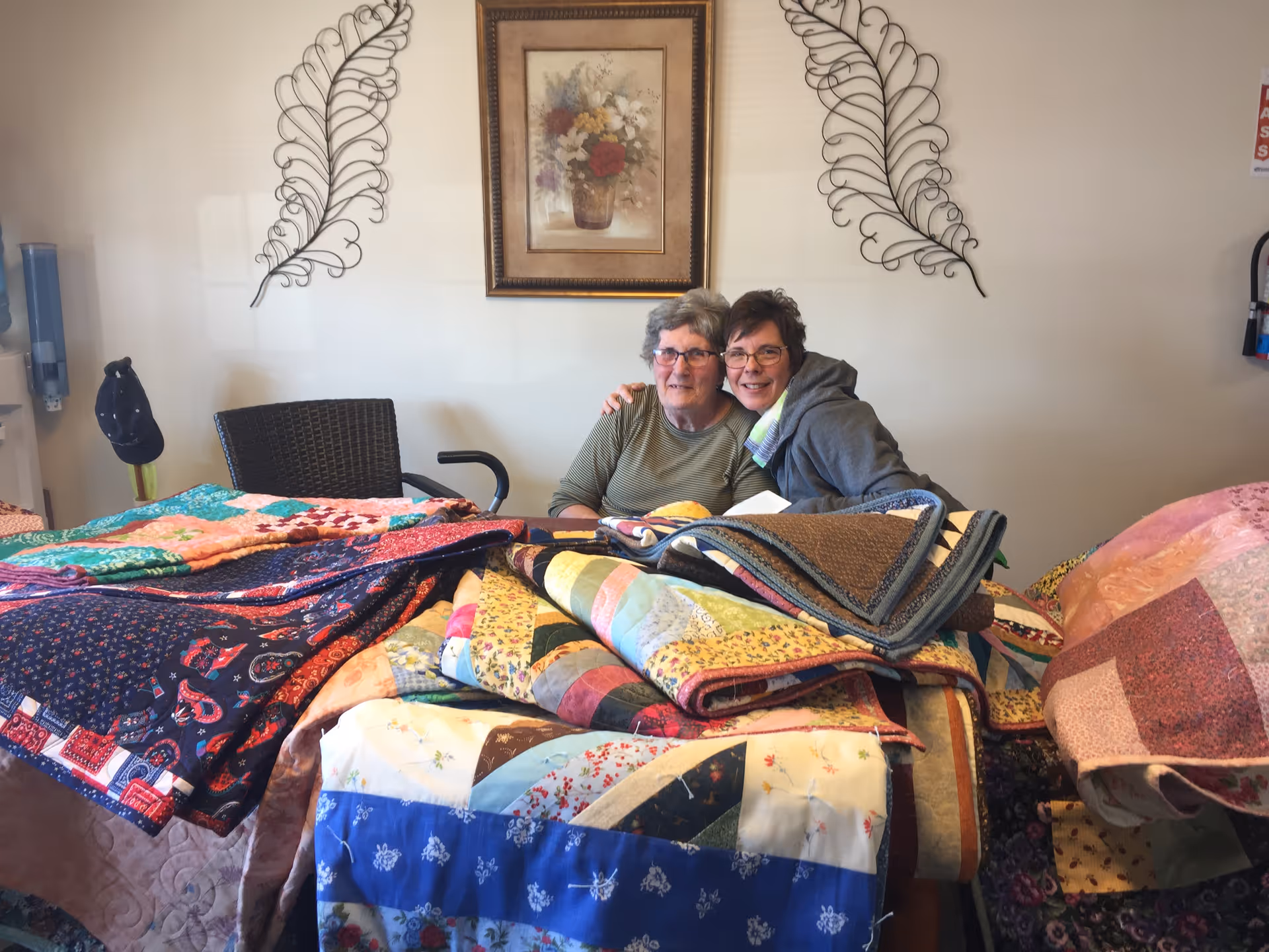 Two women are sitting together at a table covered with colorful quilts.