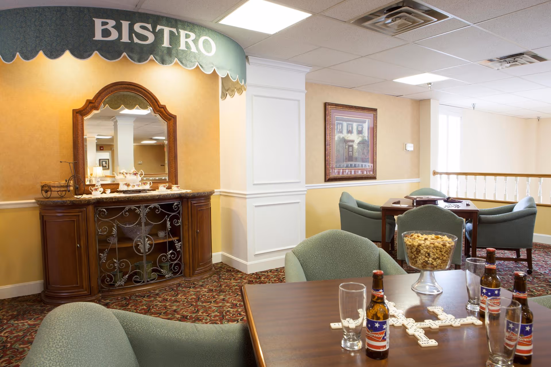 Interior view of a bistro area in a senior living facility featuring a wooden cabinet with a mirror and tea set, green upholstered chairs around tables, a bowl of mixed nuts, several bottles of beer, empty glasses, and a game of dominoes on the table. The room has patterned carpet, beige and yellow walls, and framed artwork.