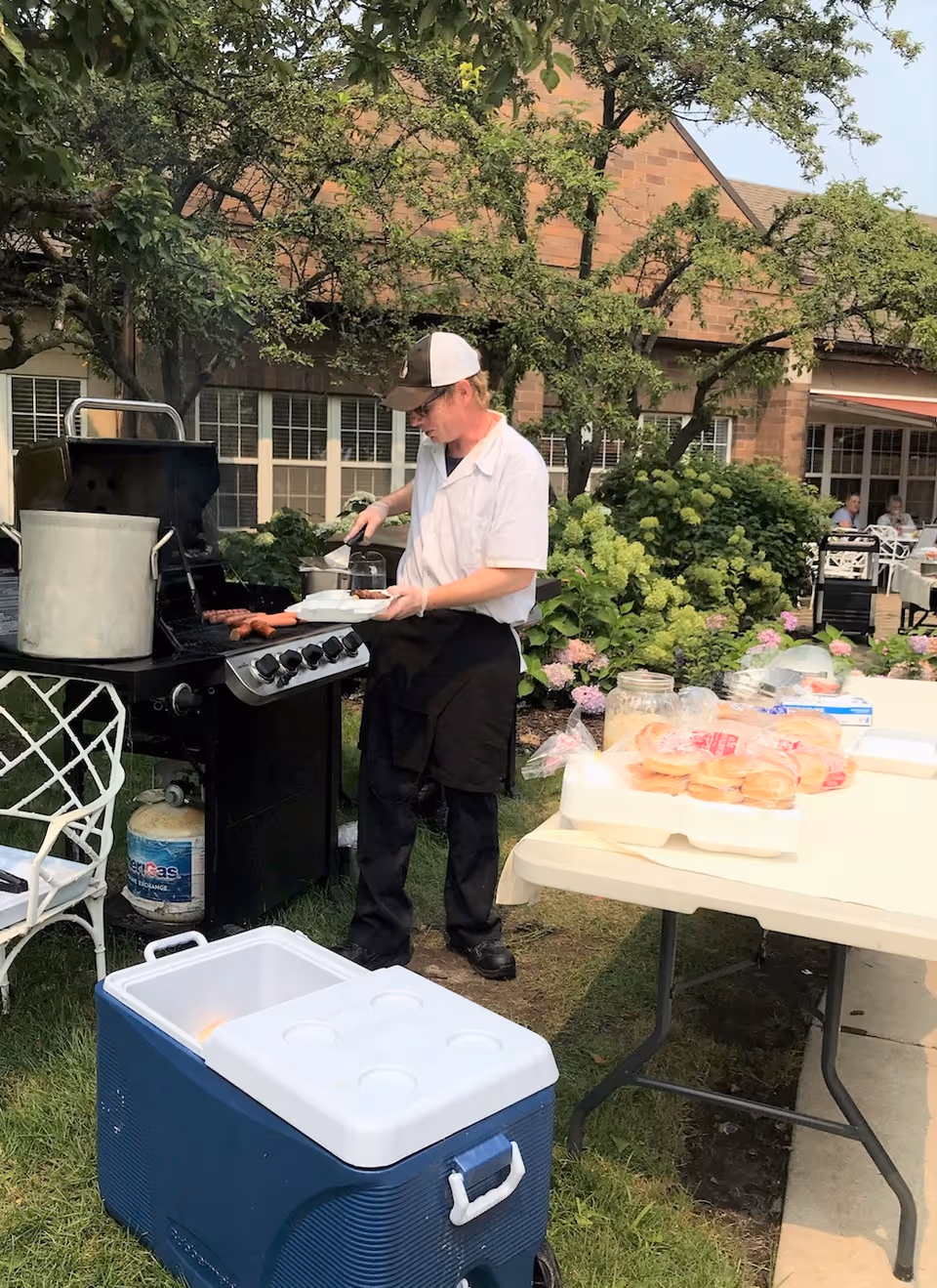 A man wearing a white shirt, black apron, and a baseball cap is grilling hot dogs on a barbecue grill outdoors near a table with buns and other food items. There is a large cooler in the foreground and a building with windows and greenery in the background.