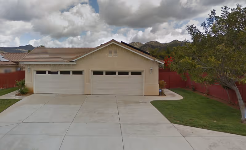 Exterior view of a single-story building with three closed garage doors, a concrete driveway, green lawn, a tree on the right, and mountains in the background under a cloudy sky.