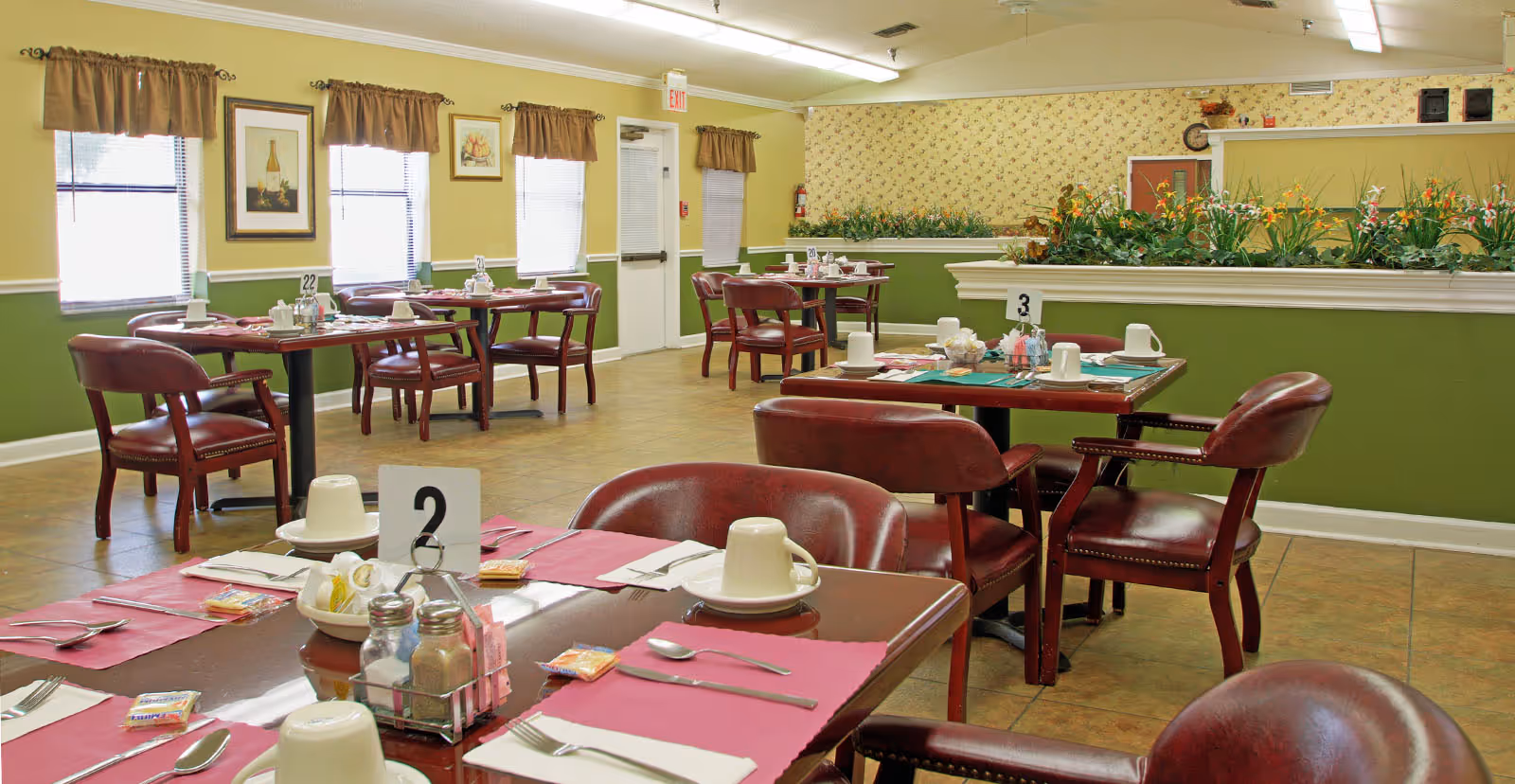 Dining room with multiple tables set with pink placemats, white cups, plates, and silverware. The room has green and yellow walls, windows with brown curtains, framed artwork, and a planter with flowers dividing the space.