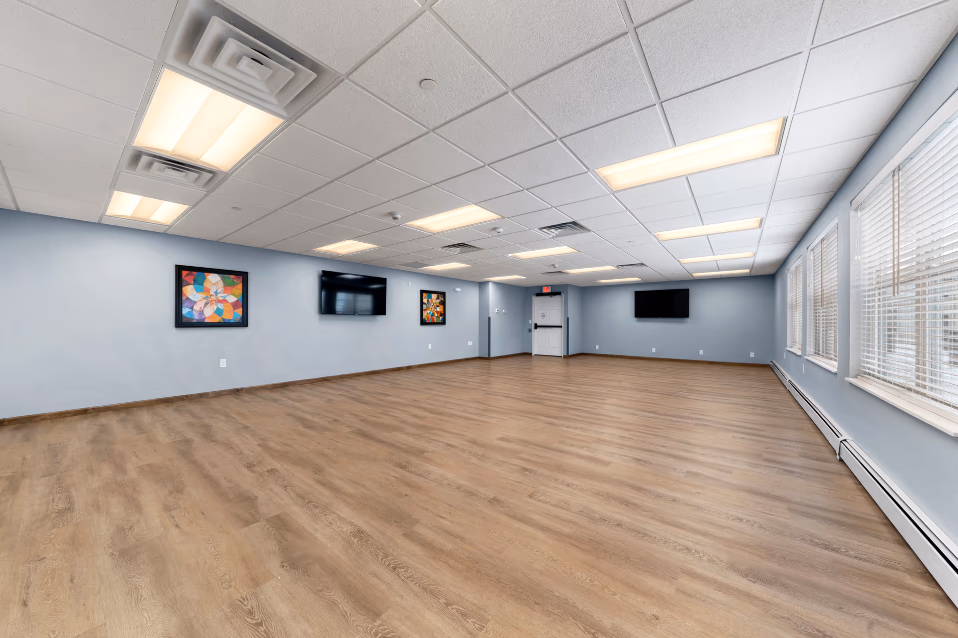 A spacious, empty room with light blue walls, wood flooring, and a white ceiling with fluorescent lights. The room features three flat-screen TVs mounted on the walls and two colorful abstract paintings. Large windows with white blinds line one side of the room, allowing natural light to enter.