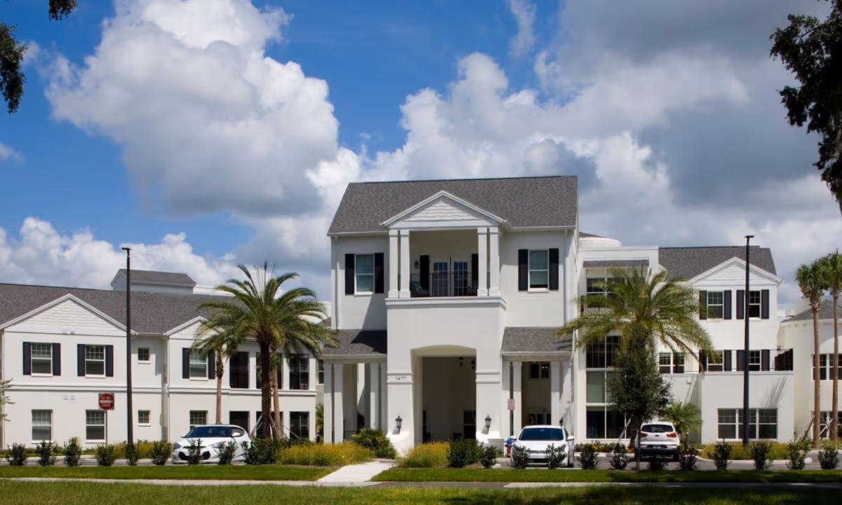Front exterior view of a large, white senior living facility building with multiple windows, black shutters, and a covered entrance. There are palm trees and parked cars in front of the building under a partly cloudy blue sky.