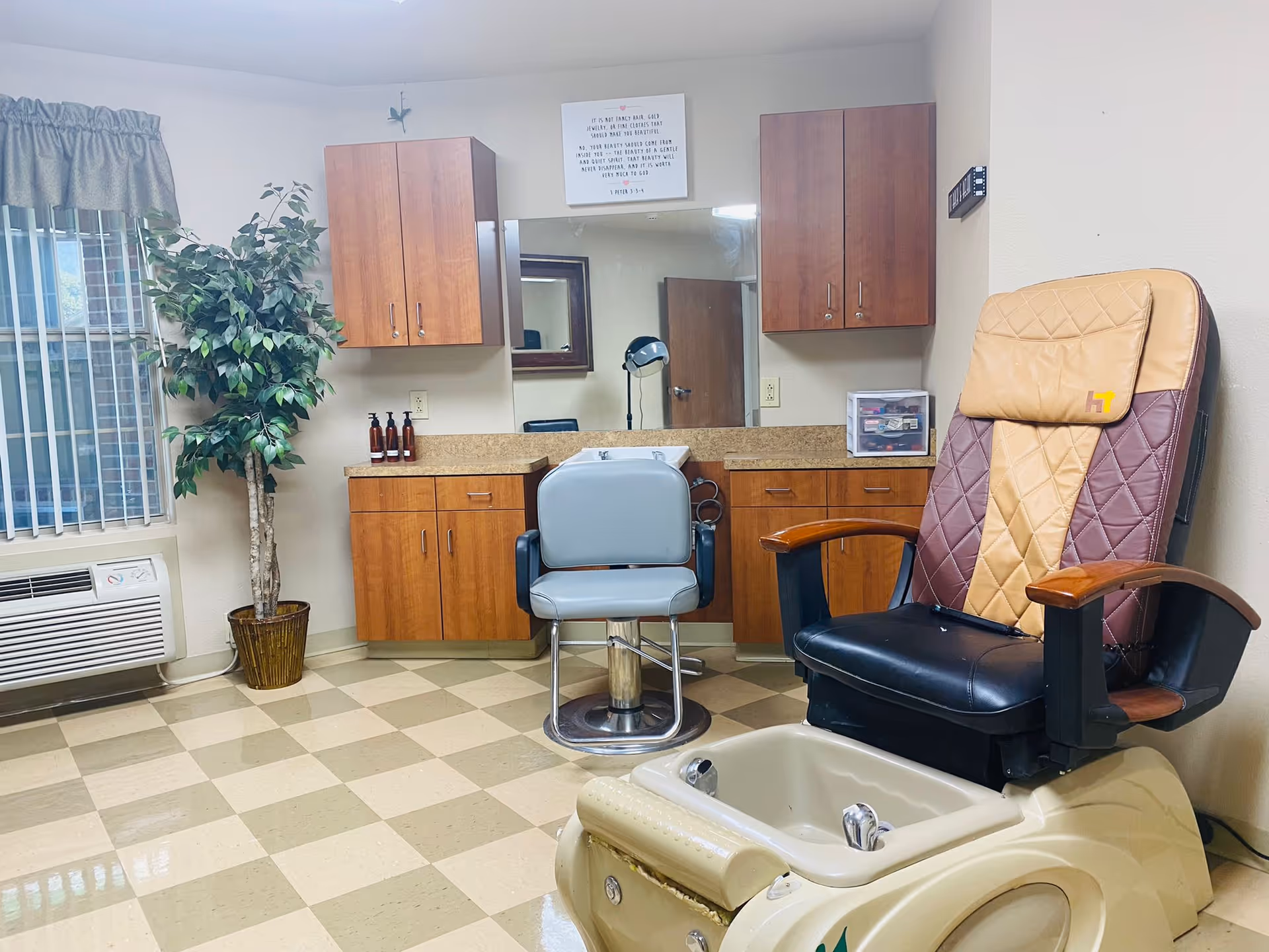 Interior view of a salon area in a healthcare facility featuring a pedicure chair with tan and brown upholstery, a light blue salon chair, wooden cabinets with a granite countertop, a large mirror, a potted plant near a window with vertical blinds, and a wall-mounted air conditioning unit.