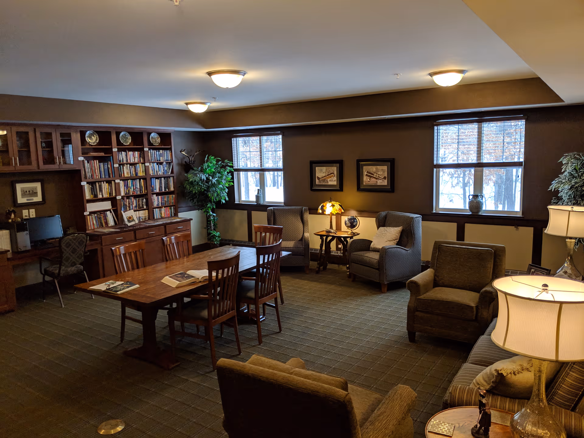 A cozy senior living common room with bookshelves, a wooden table and chairs, armchairs, lamps, and windows showing snow outside.
