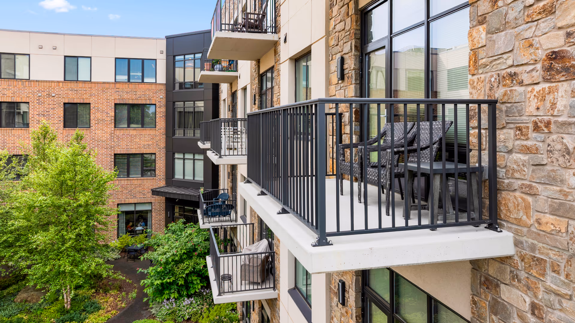 View of multiple balconies on a senior living facility building with stone and brick exterior walls, overlooking a landscaped courtyard with trees and greenery.