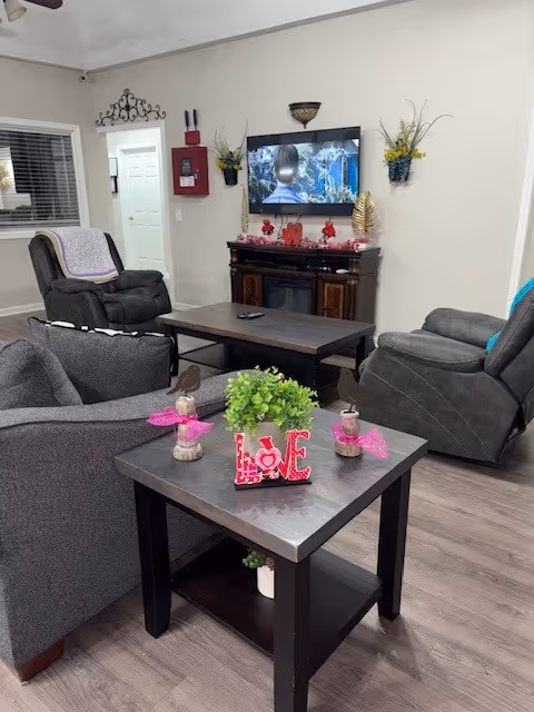 A cozy living room with three gray recliner chairs arranged around a dark wooden coffee table and an end table. The end table has decorative items including a green plant and a pink 'LOVE' sign. A flat-screen TV is mounted on the wall above a fireplace with red and gold decorations. The walls are light-colored with two wall-mounted flower pots and a window with blinds on the left side.