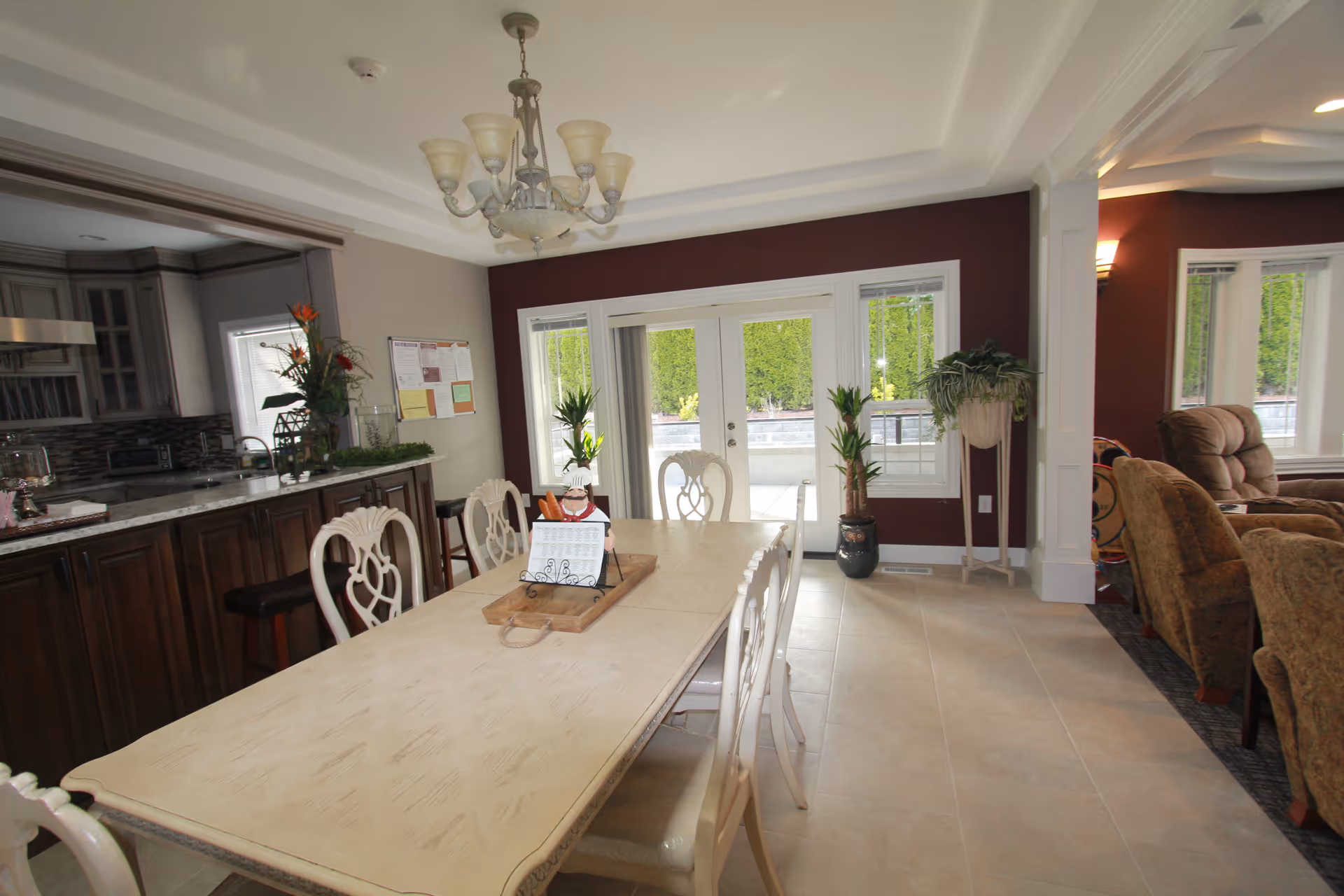 A bright dining area with a long wooden table surrounded by white chairs. The room features a chandelier hanging from the ceiling, large windows and glass doors letting in natural light, and a kitchen area with dark wooden cabinets and a granite countertop. There are plants placed near the windows and on the countertop, and a cozy living area with upholstered chairs is visible to the right.