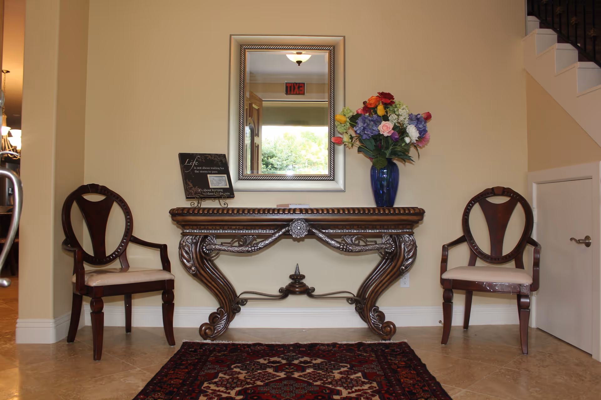Entryway interior with an ornate wooden console table beneath a mirror, a vase of flowers, two chairs, and a decorative rug.