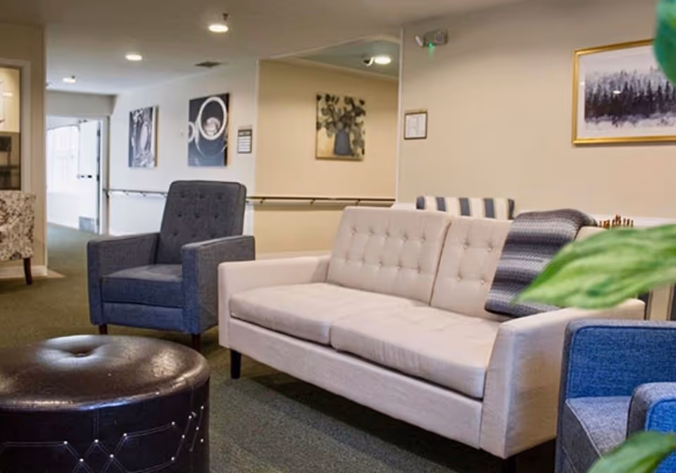 A cozy seating area in a senior living facility with a light beige tufted sofa, a dark blue armchair, a round black leather ottoman, and a striped throw blanket on the sofa. The walls are decorated with framed artwork, and there is a green plant partially visible in the foreground.