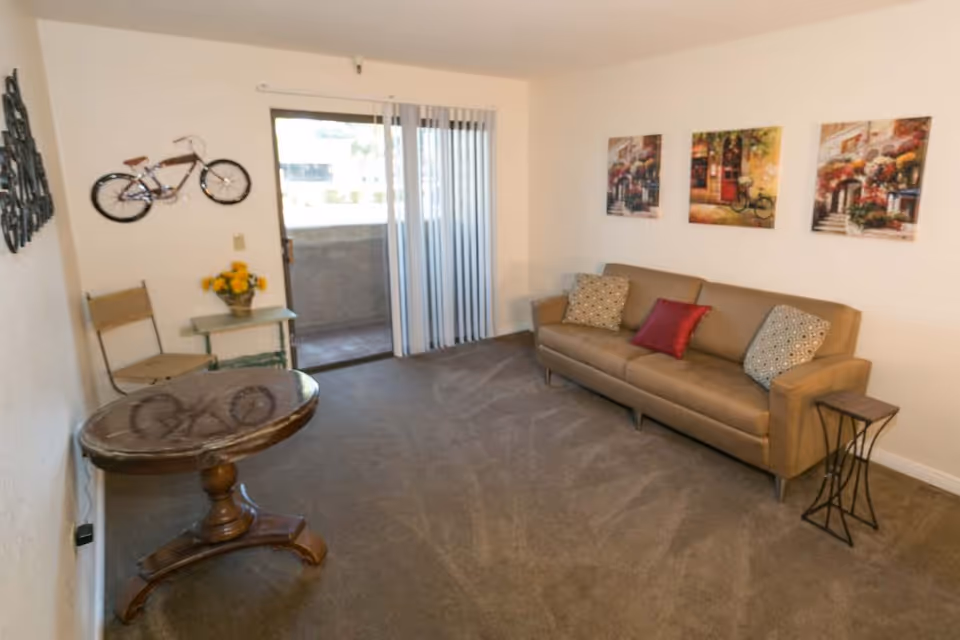 A living room with a brown leather sofa adorned with three decorative pillows, a small side table, a round wooden table, a chair, and a small table with a vase of yellow flowers. There are three colorful paintings on the wall above the sofa, a decorative bicycle mounted on the wall, and a sliding glass door with vertical blinds leading to an outdoor balcony.