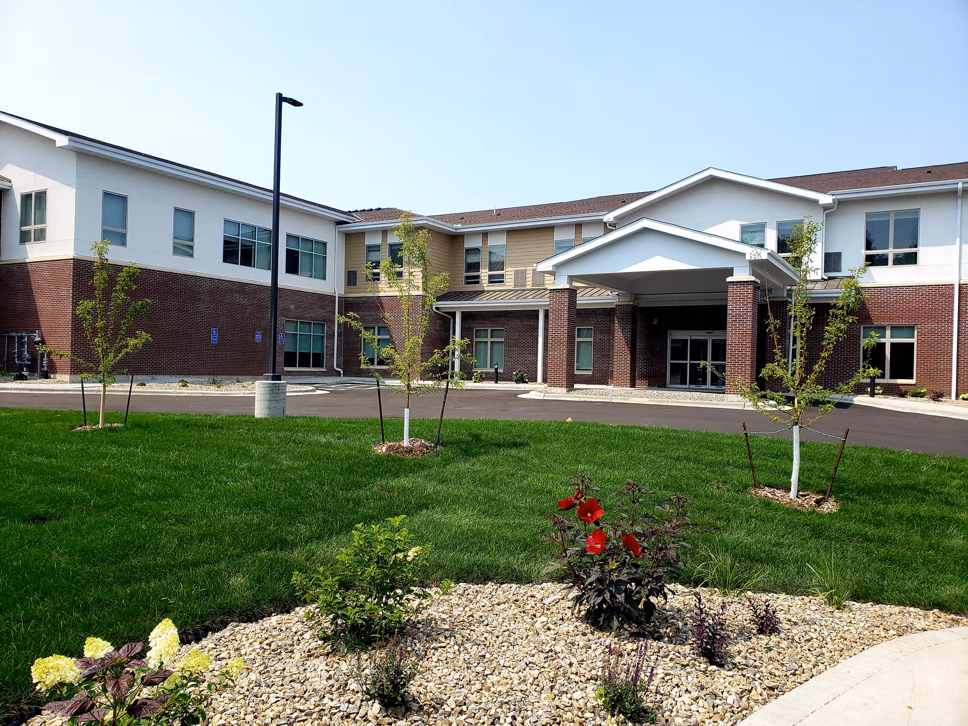 Front entrance of a two-story senior living building with a covered porte-cochere, landscaped lawn and young trees.