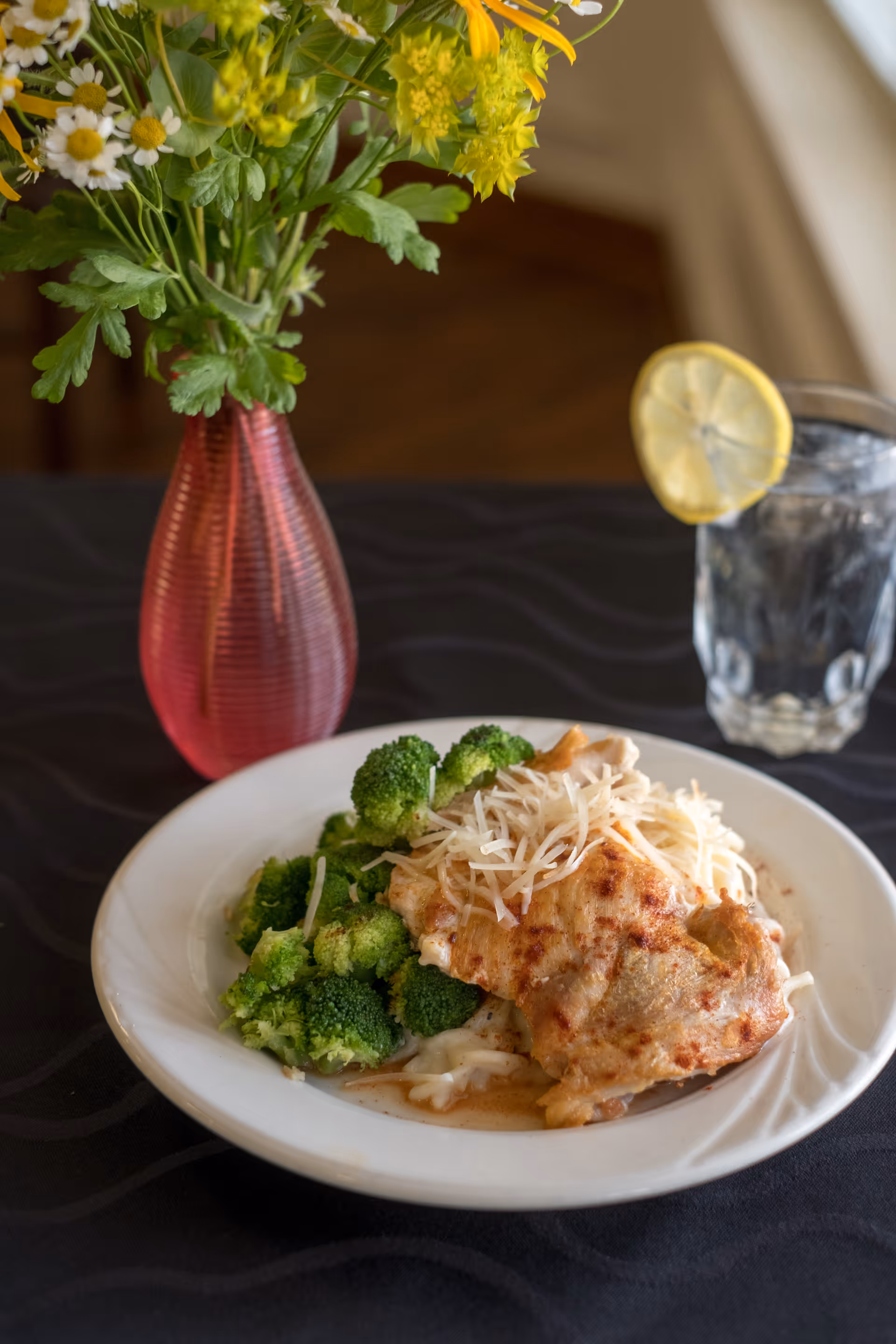 A plate of food featuring cooked broccoli and a serving of chicken topped with shredded cheese, placed on a black tablecloth. Next to the plate is a glass of water with a lemon slice on the rim and a red vase with yellow and white flowers in the background.