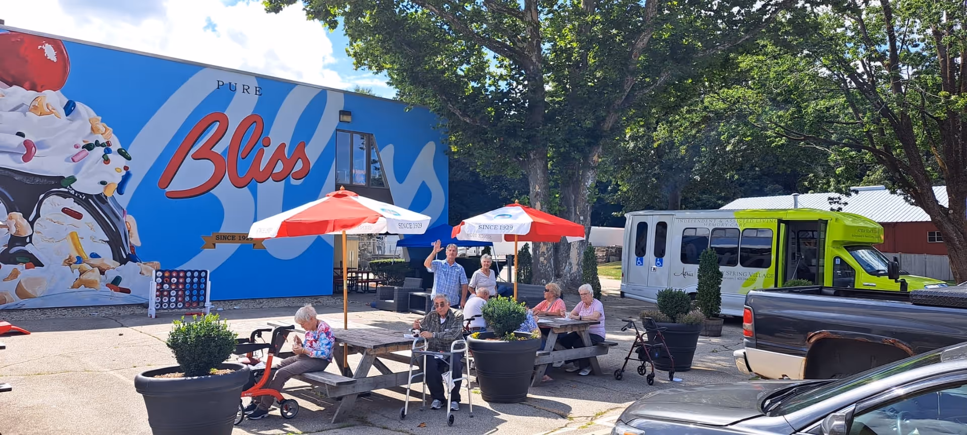Outdoor patio with seniors seated at picnic tables under red umbrellas in front of a large blue mural that reads "Bliss" and a green facility shuttle nearby.