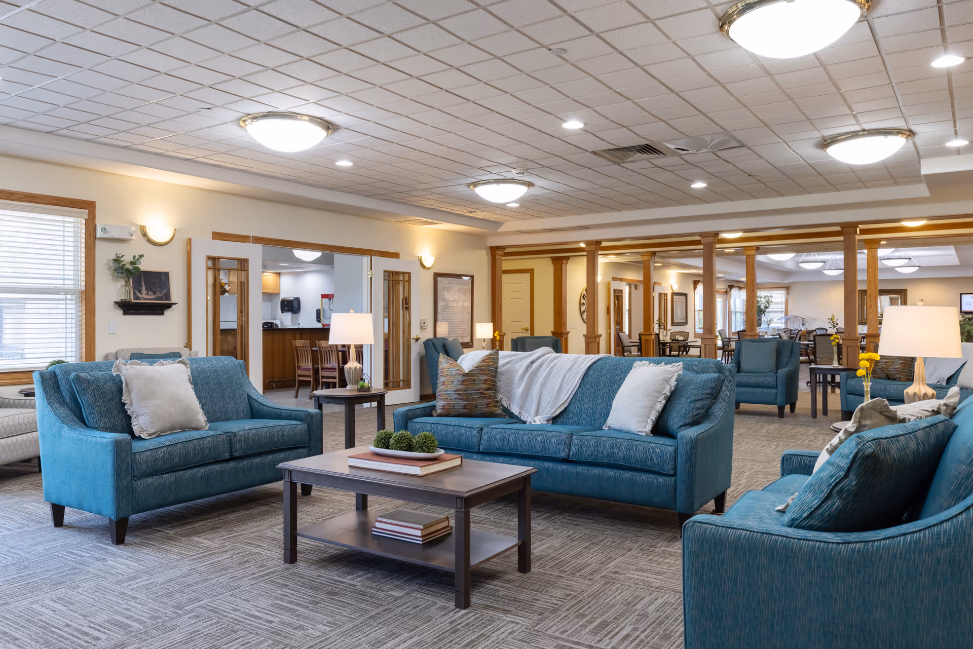 A spacious and well-lit living room area in a senior living facility with teal upholstered sofas and armchairs arranged around a wooden coffee table. The room features a carpeted floor, multiple ceiling lights, and large windows with blinds. In the background, there are wooden columns separating the living area from a dining space with tables and chairs.