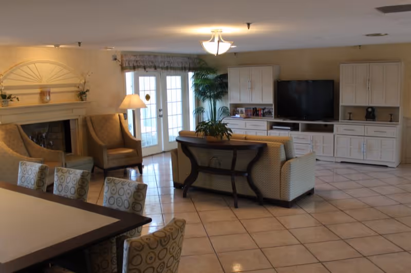 A spacious living room area with tiled floors, a beige sofa with a wooden table behind it, two armchairs near a fireplace, a floor lamp, a large TV on a white entertainment center, and French doors leading outside. There are also some plants and a dining table with patterned chairs in the foreground.