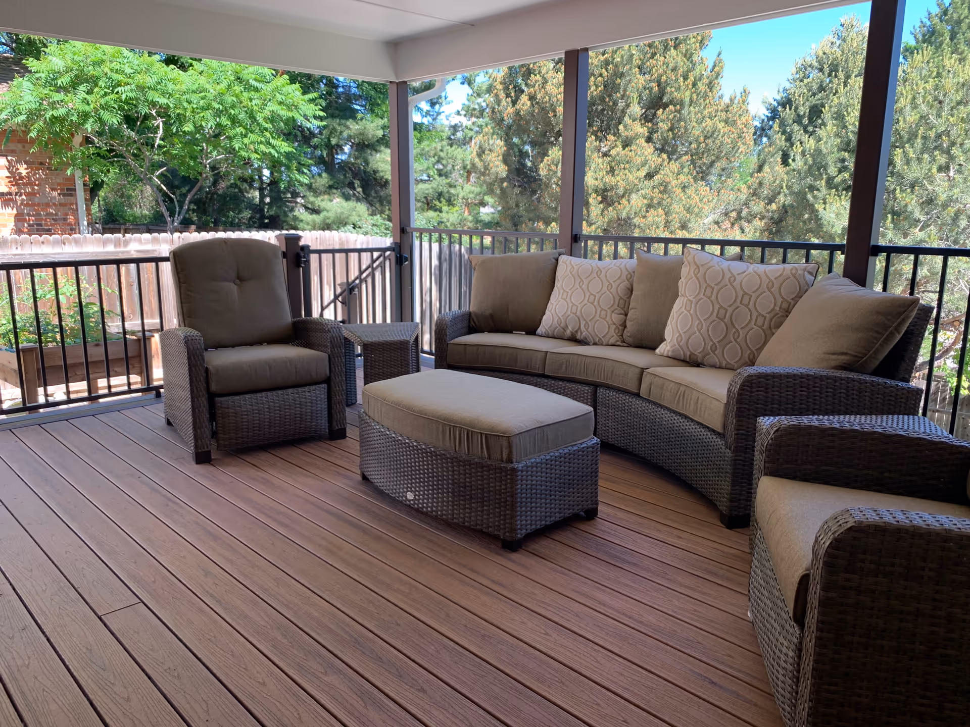 A covered outdoor patio area with brown wicker furniture including a curved sofa with beige cushions and patterned pillows, two armchairs, and an ottoman. The patio has wooden flooring and is surrounded by a black metal railing. Trees and greenery are visible outside the patio.