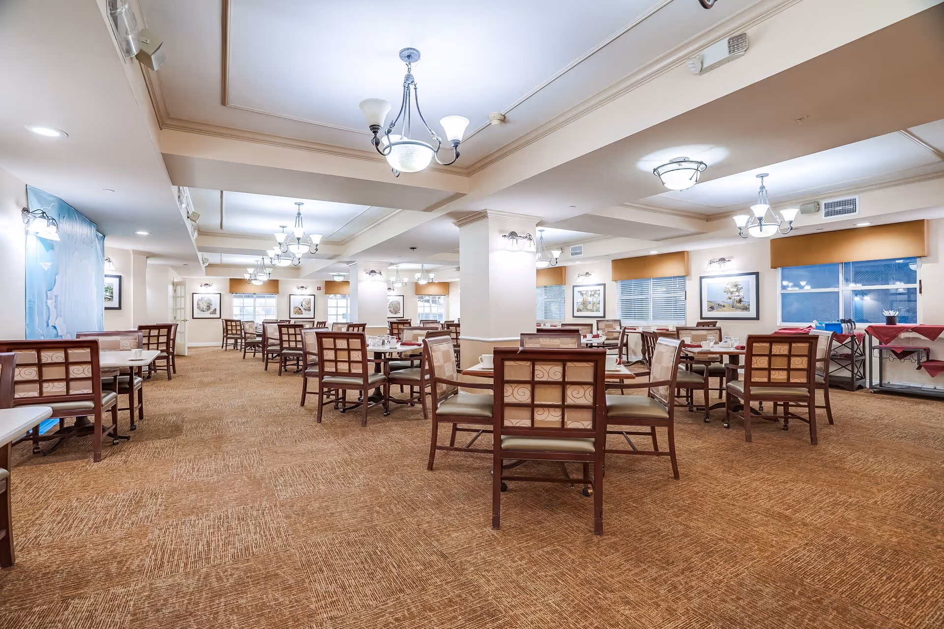 Spacious dining room in a senior living facility with multiple wooden tables and chairs arranged neatly. The room is well-lit with ceiling lights and wall sconces, featuring carpeted floors and large windows with blinds. Artwork is displayed on the walls, and there are tables with red tablecloths along the right side.